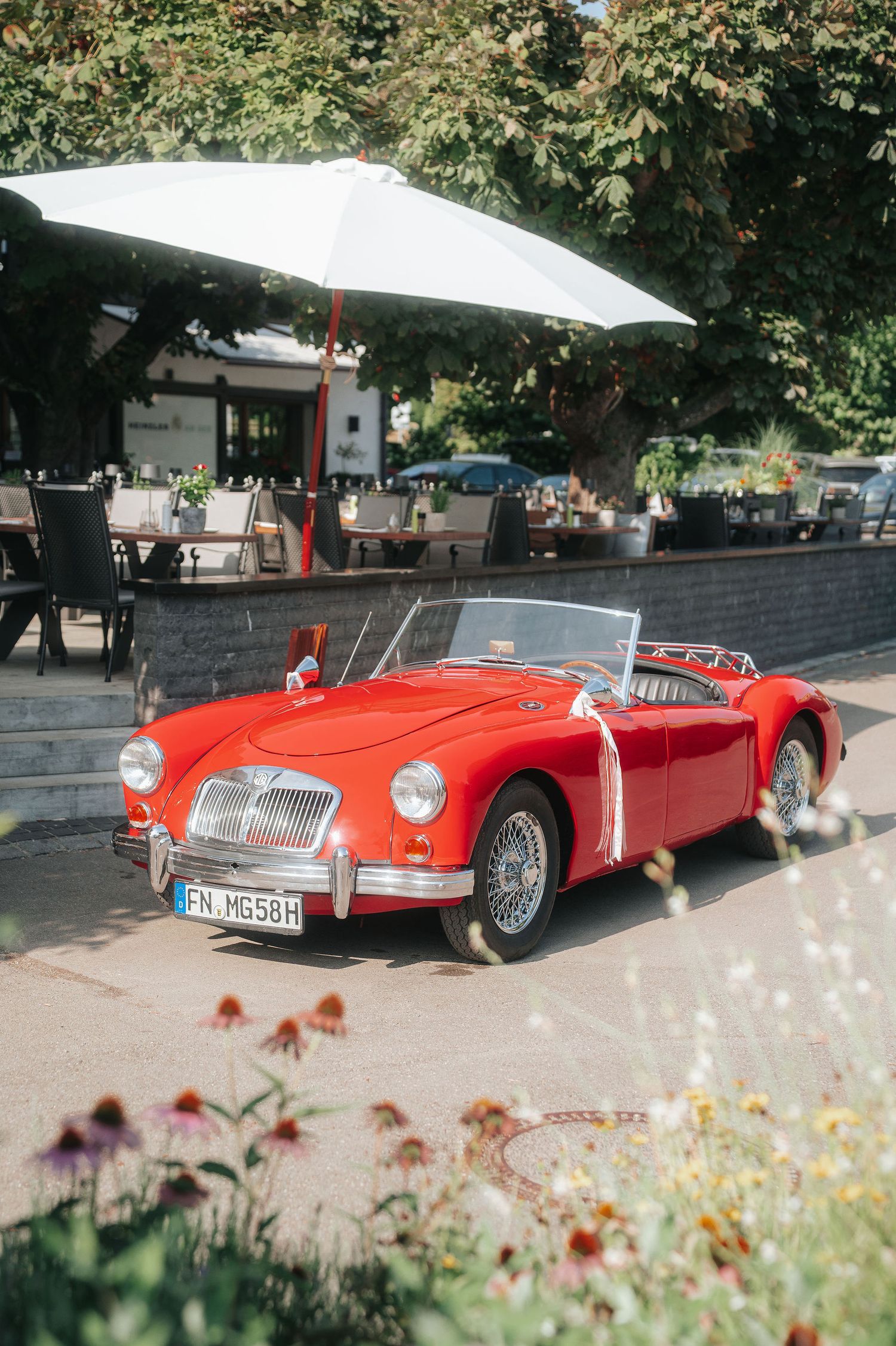 Red vintage MG roadster convertible parked beside an outdoor café with a white umbrella, wildflowers in the foreground.