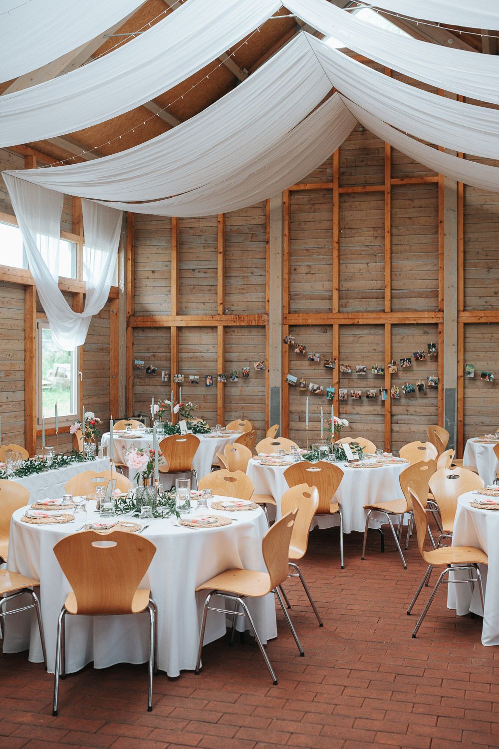 Wedding reception venue decorated with white drapes and string lights features wooden chairs and round tables on brick floor.