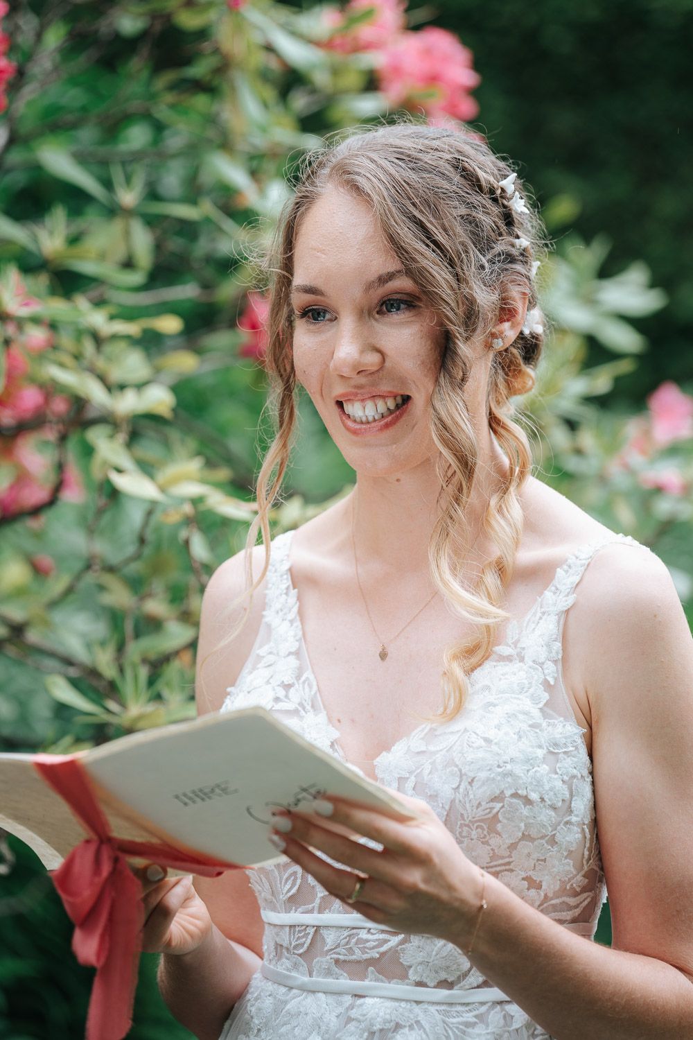 A bride in a white lace wedding dress smiles while holding a book in a garden with pink flowers.