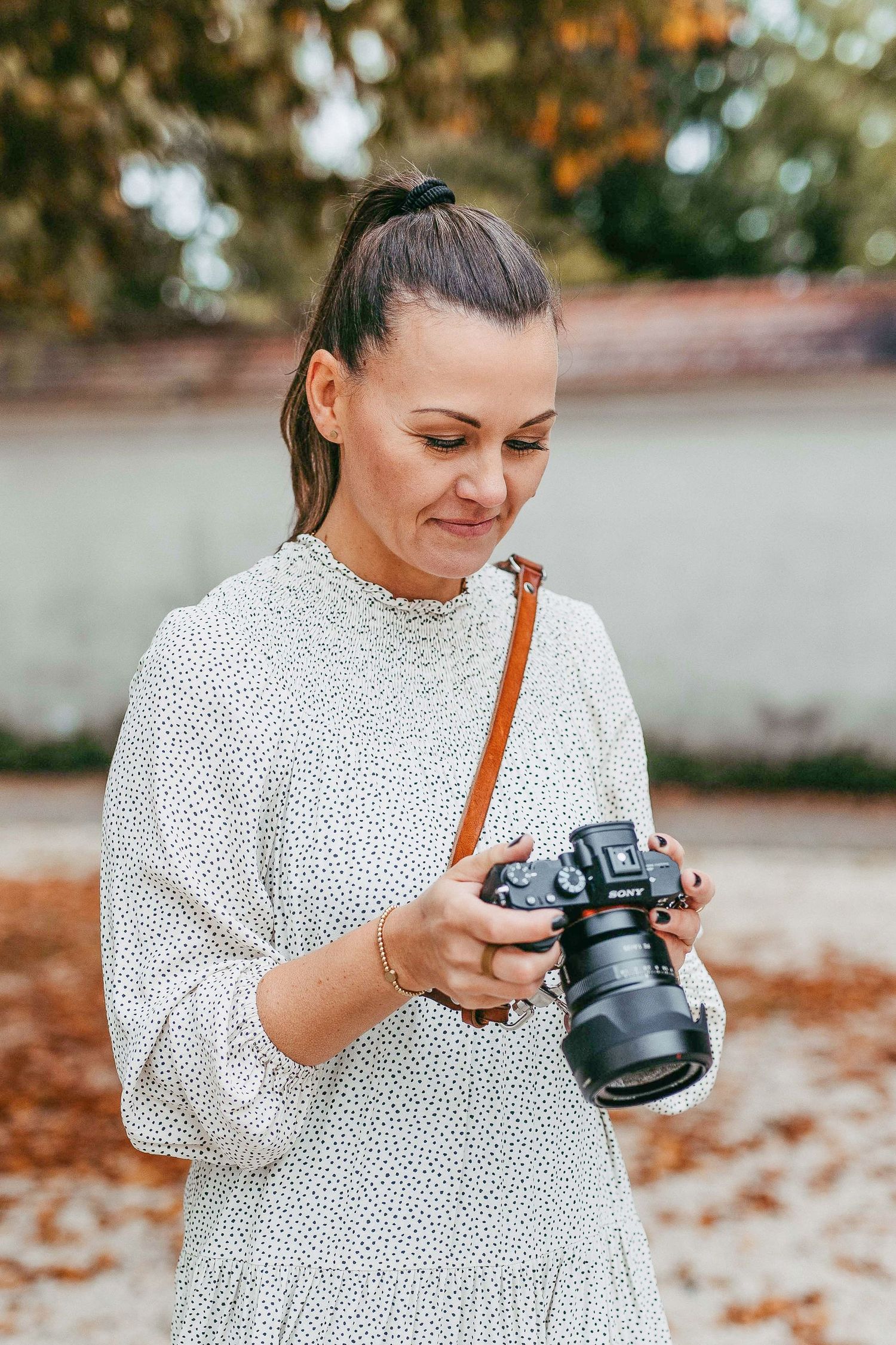 A photographer in a white polka dot blouse examines settings on a professional DSLR camera outdoors during autumn.