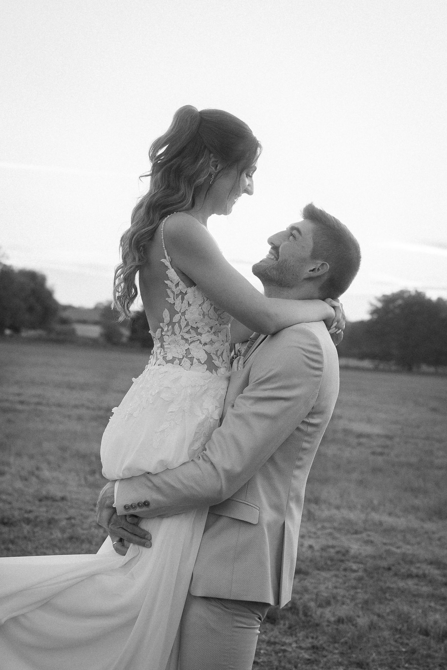 Black and white wedding photo of a groom lifting his smiling bride in a lace gown in an open field at sunset.