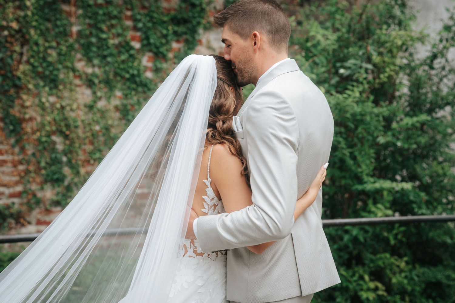 Groom in a light gray suit embraces a bride in a lace wedding dress and long veil beside a green ivy-covered brick wall.