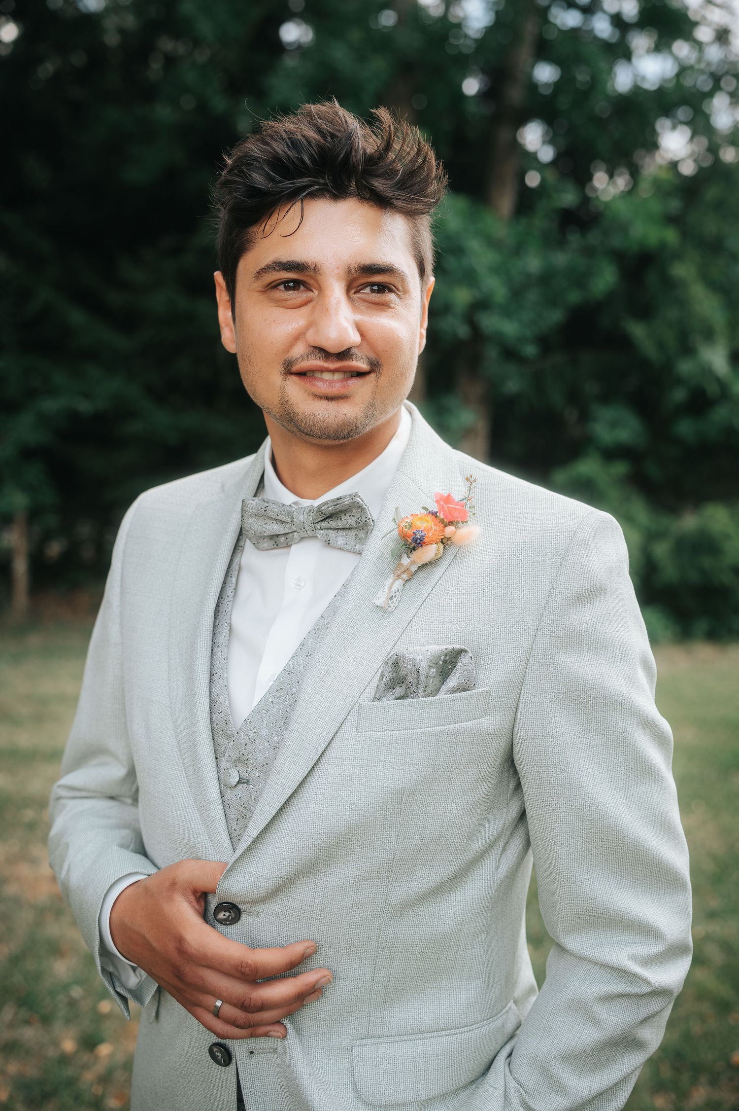 Smiling groom in a light gray suit with matching vest, bow tie, pocket square and floral boutonniere stands outdoors in a green park.