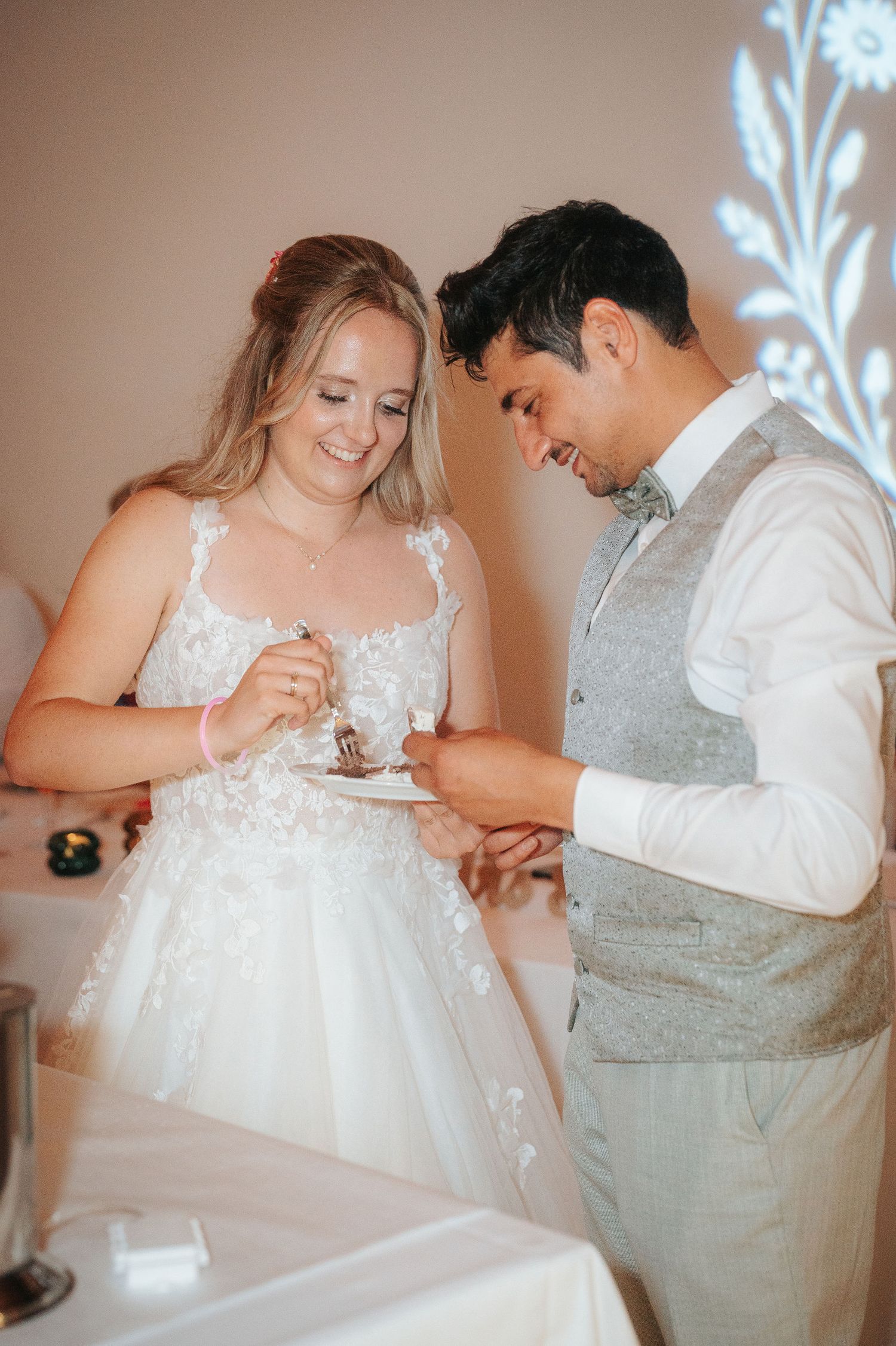 A smiling bride in a lace wedding gown and a groom in a gray vest share a bite of wedding cake at their reception.