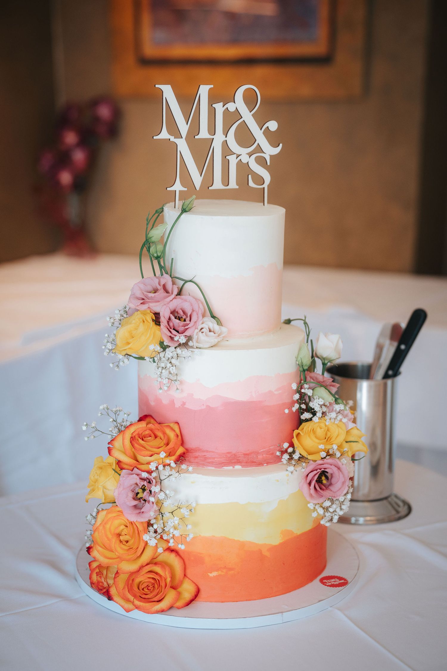 Three-tier ombre wedding cake in pink, coral and yellow with fresh roses, baby's breath and a Mr & Mrs cake topper.