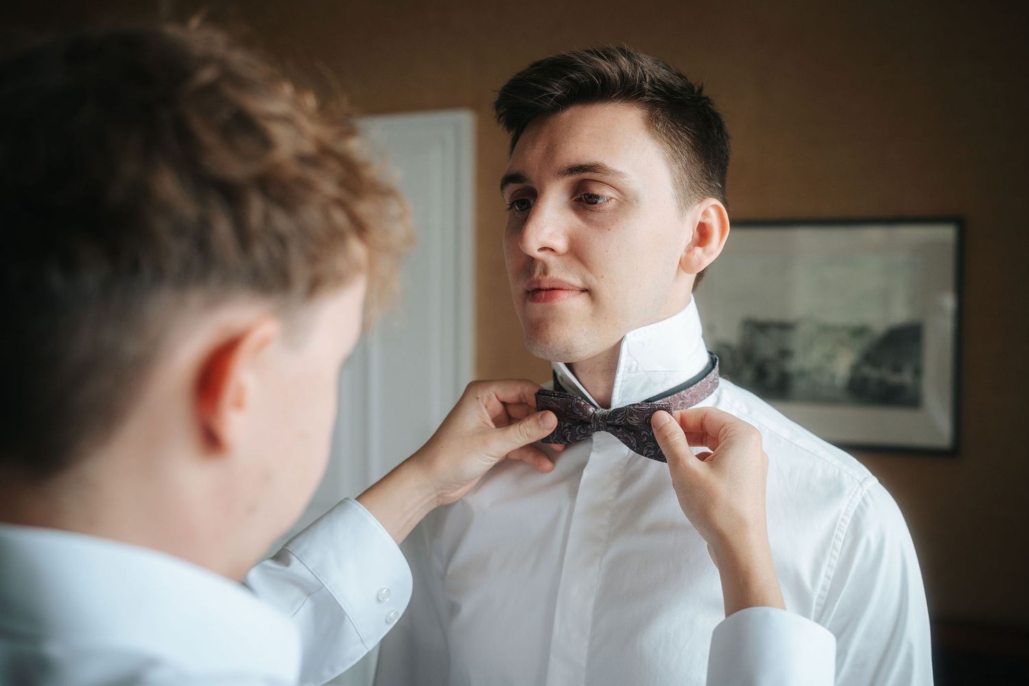 A groomsman in a white dress shirt helps a smiling man adjust his gray bow tie while getting ready for a wedding in a warmly lit room.