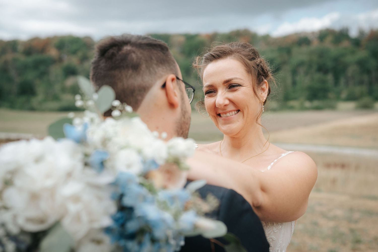 Smiling bride in a white dress embraces her groom beside a bouquet of white and blue flowers in a scenic outdoor wedding photo.