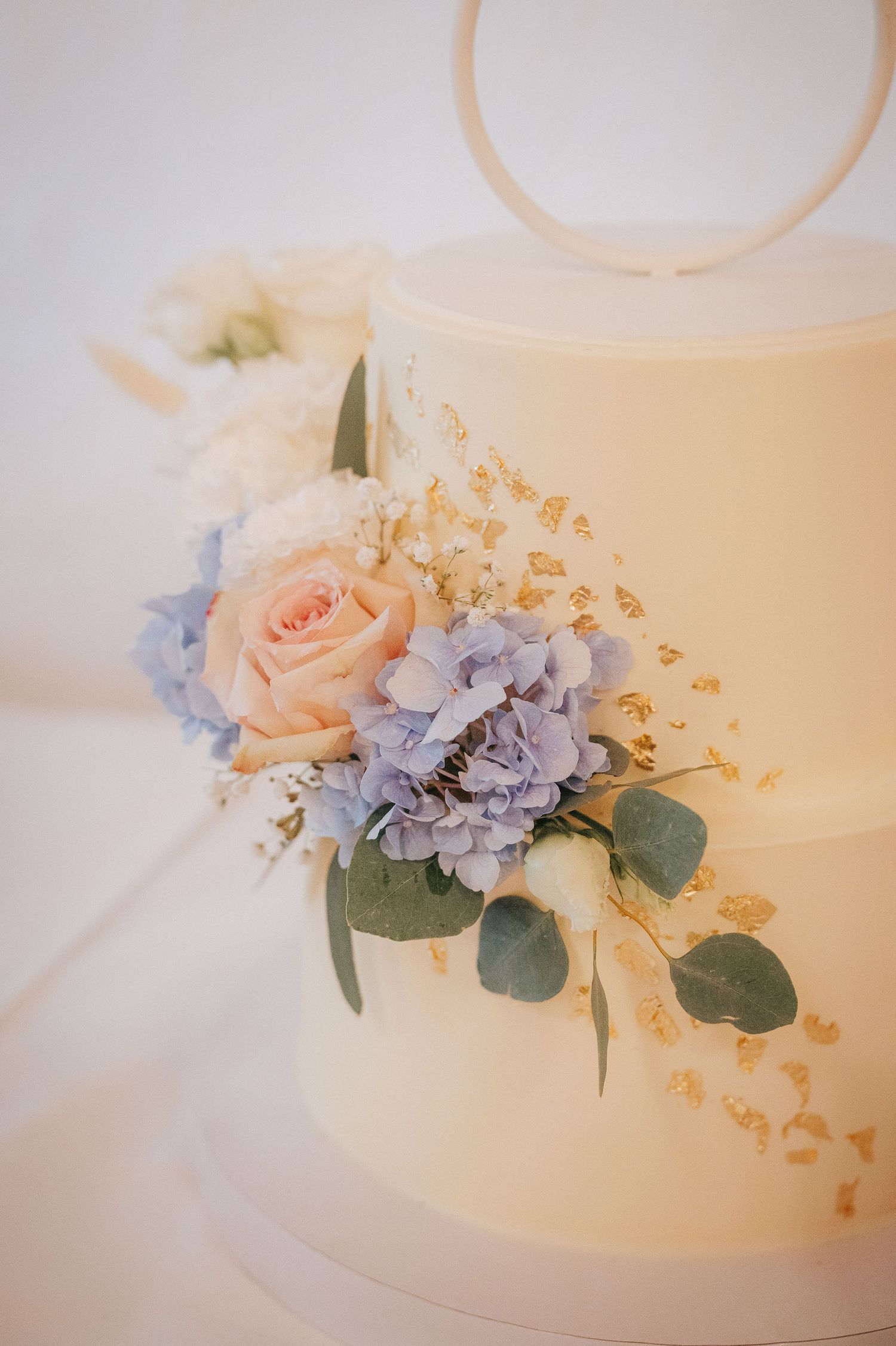 Two-tier ivory wedding cake decorated with blue hydrangea, a peach rose, white blooms, eucalyptus leaves and gold leaf flakes.