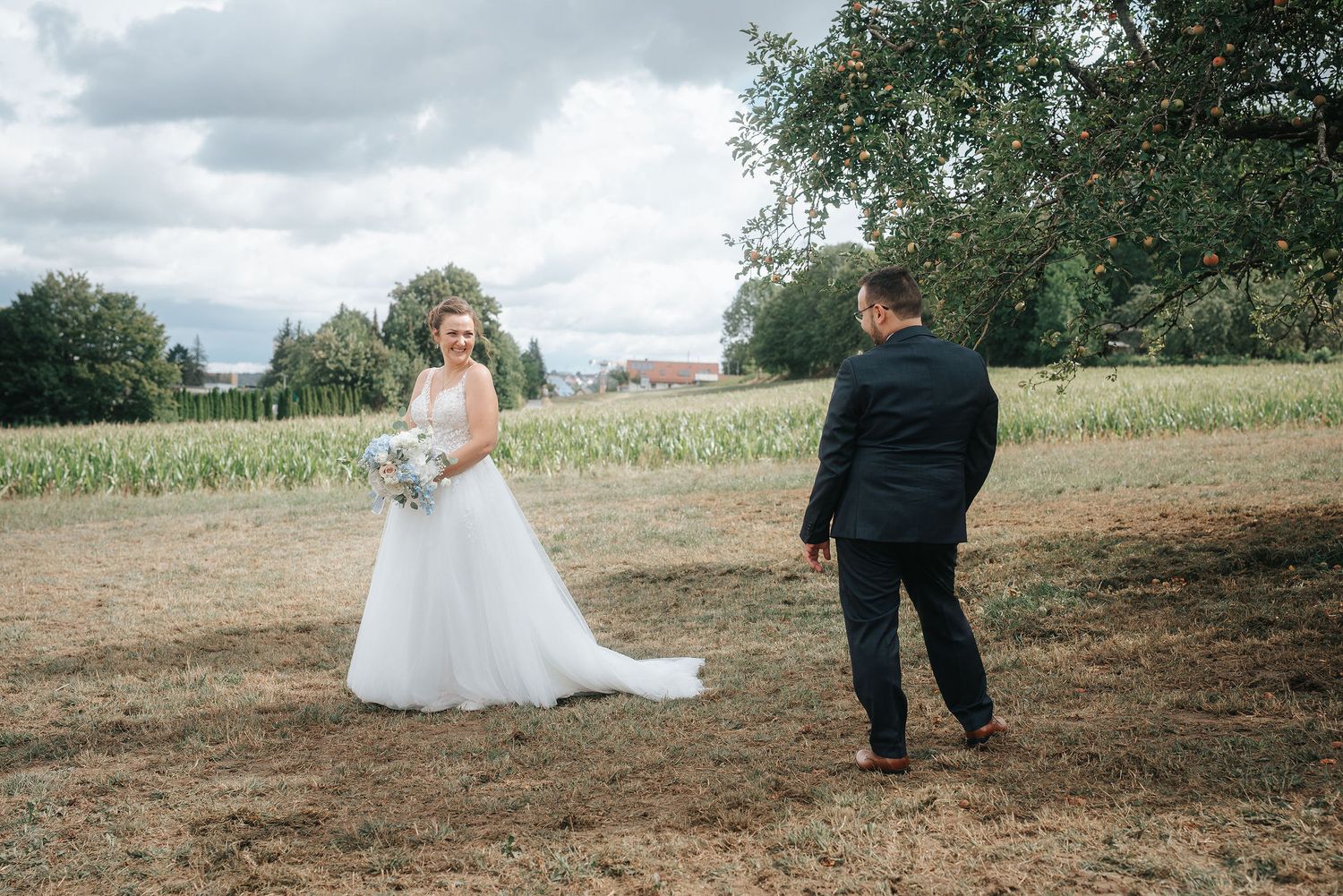 Bride in a white wedding gown holding a bouquet faces a groom in a dark suit in a grassy field with trees and a cloudy sky.
