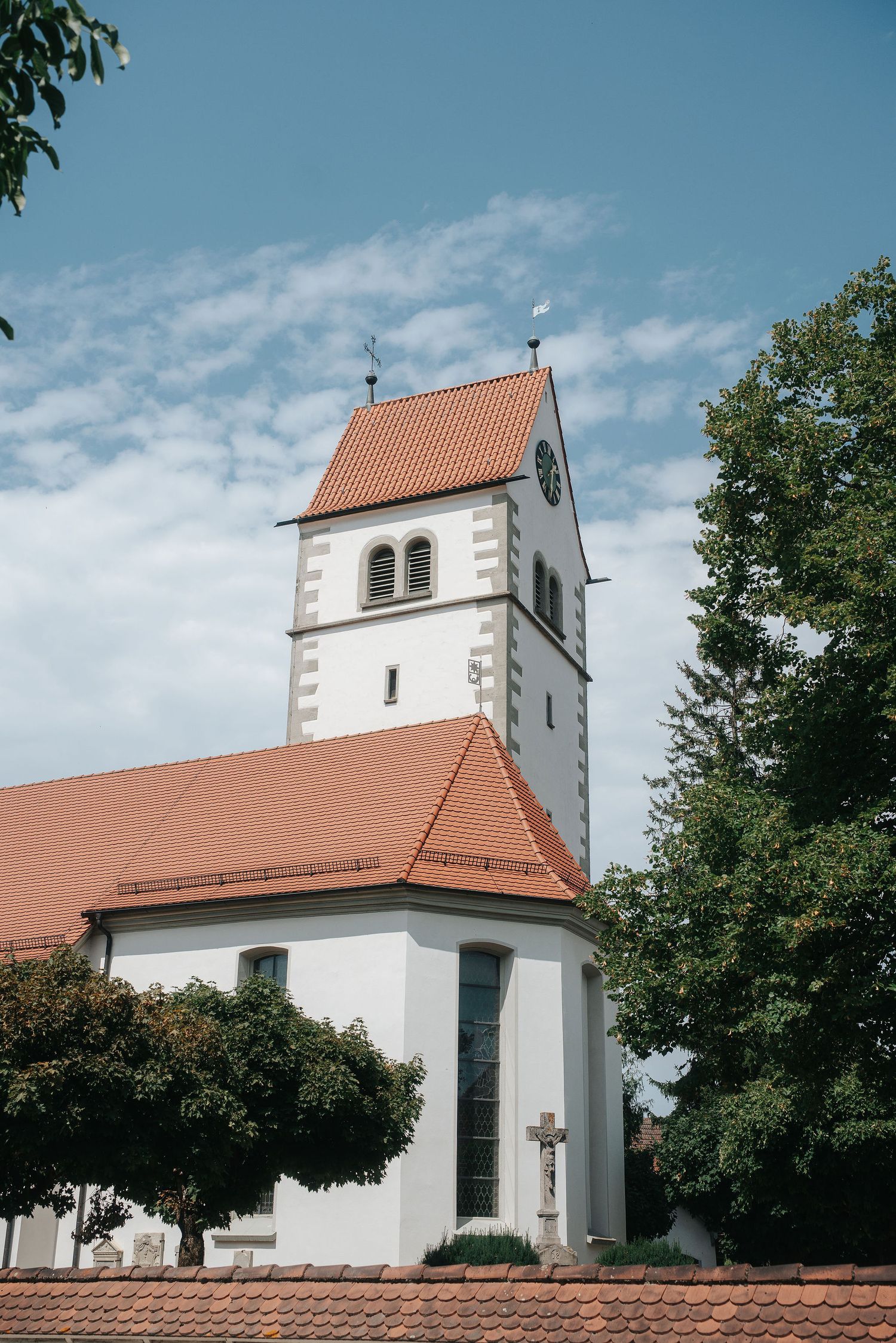 White village church with a clock tower and orange tiled roof, a stone crucifix and green trees beneath a blue sky with soft clouds.