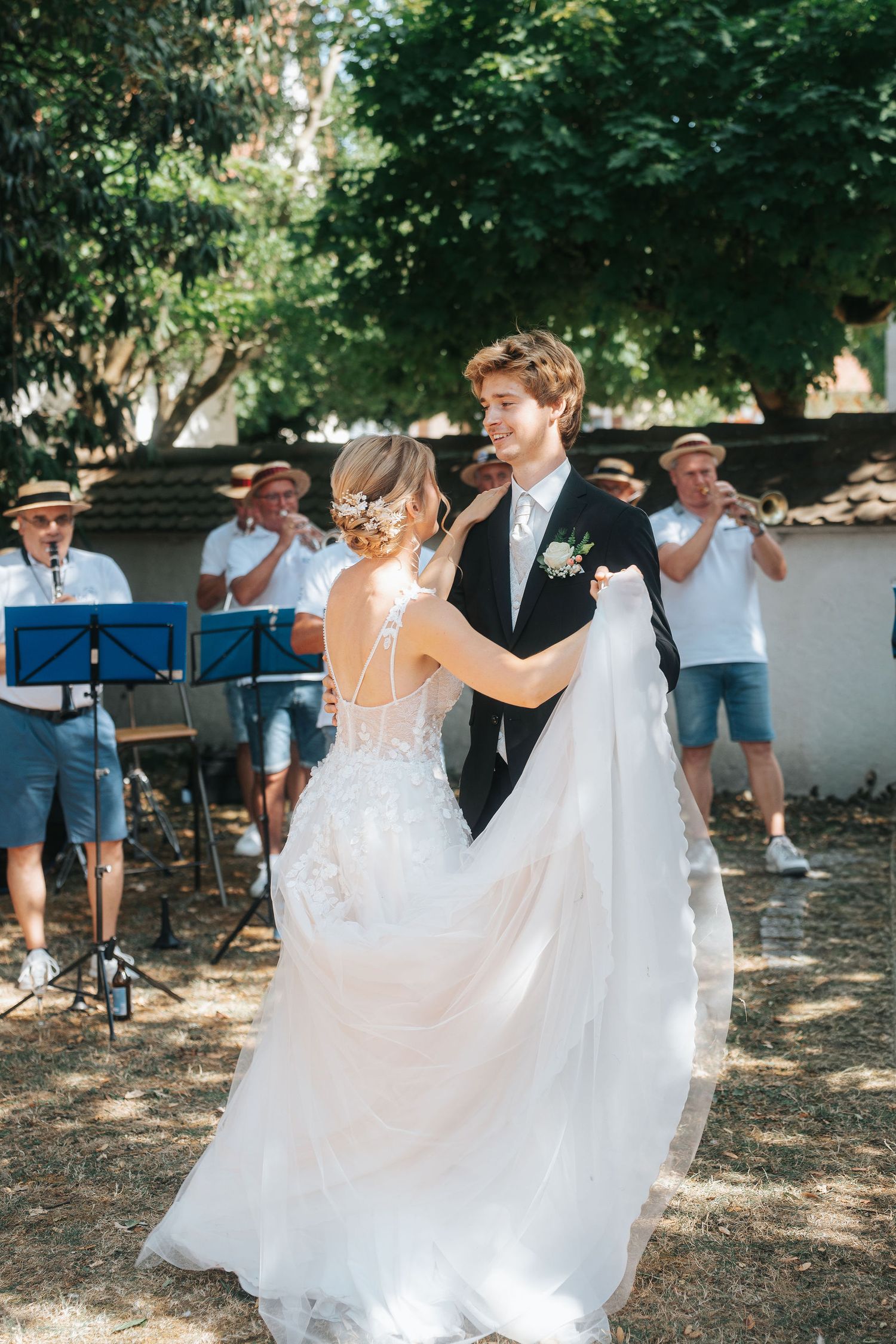 Bride in a flowing white lace gown shares a first dance with a groom in a dark suit as musicians in straw hats play outdoors.