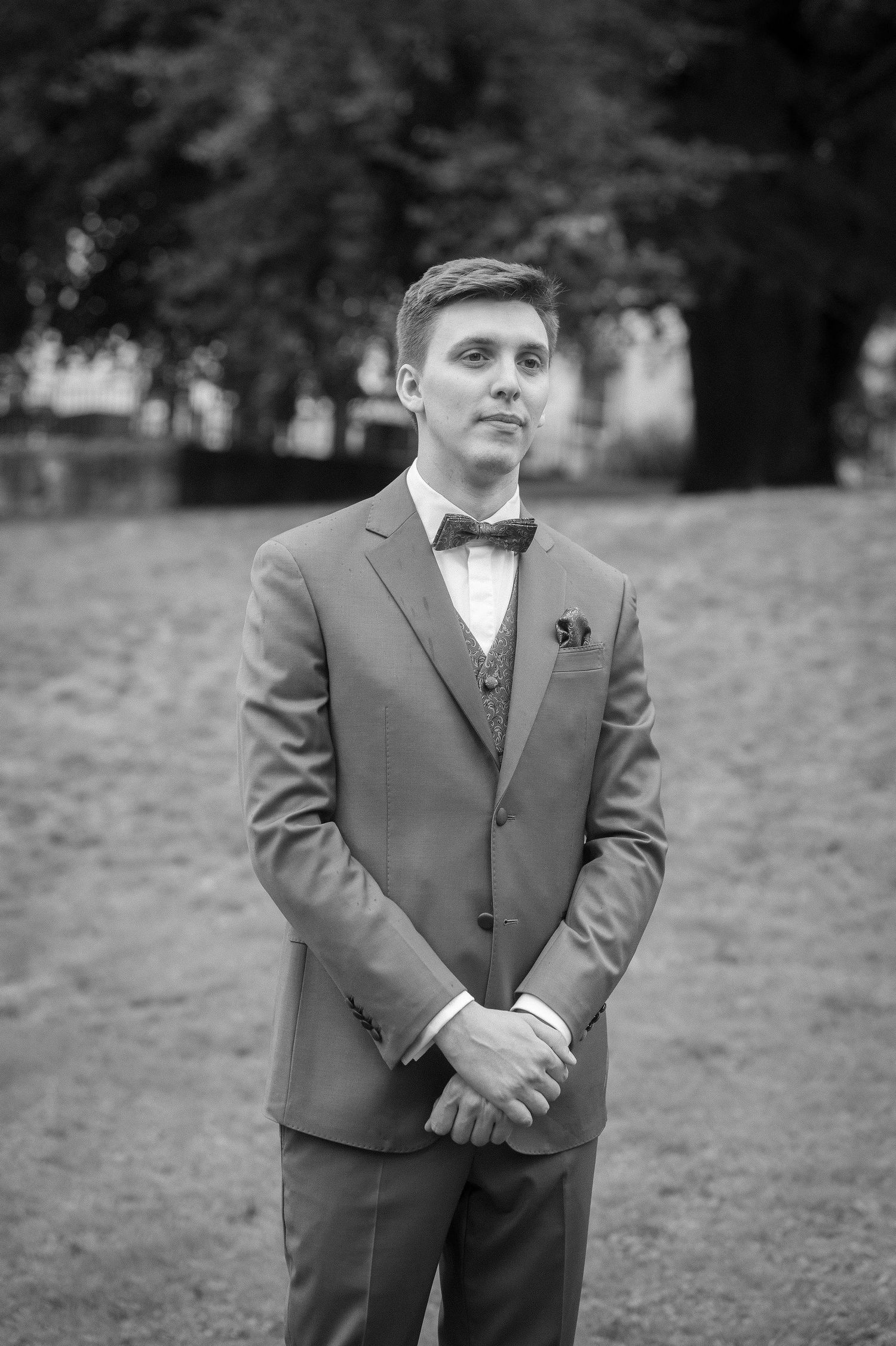 Black and white portrait of a young man in a tailored suit, vest and bow tie standing with clasped hands in a park.
