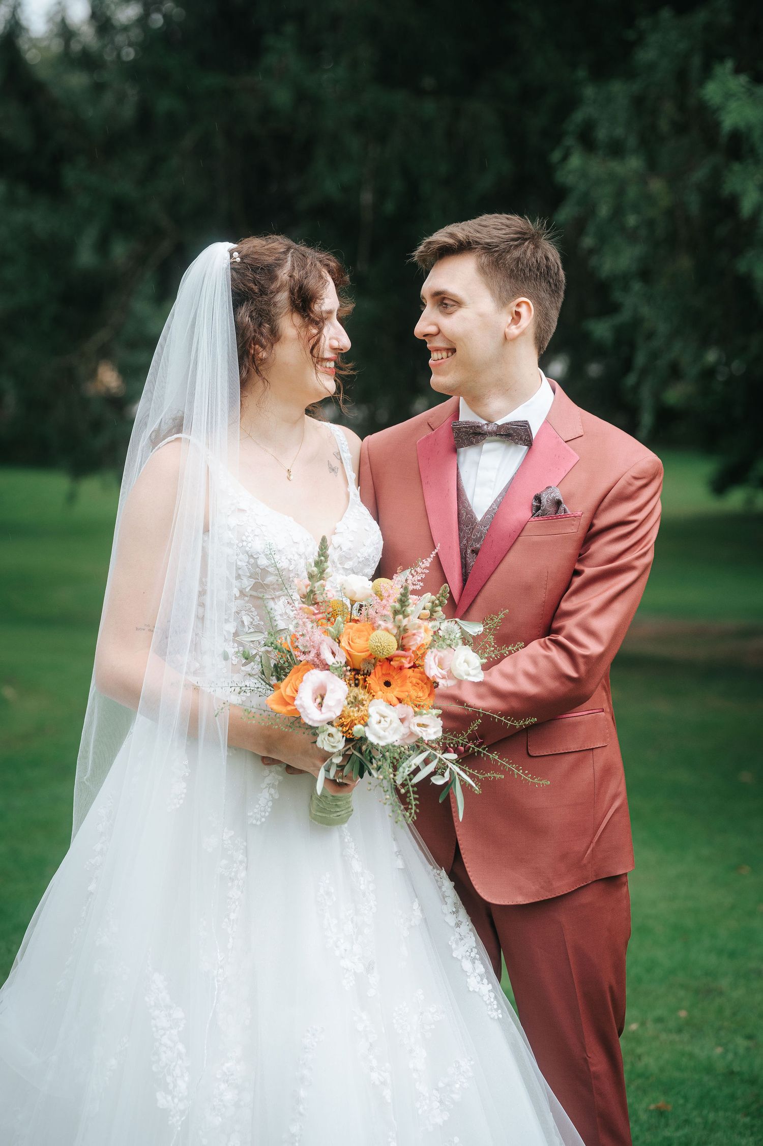 A smiling bride in a lace gown and veil gazes at her groom in a rust suit while holding an orange and white bouquet outdoors.
