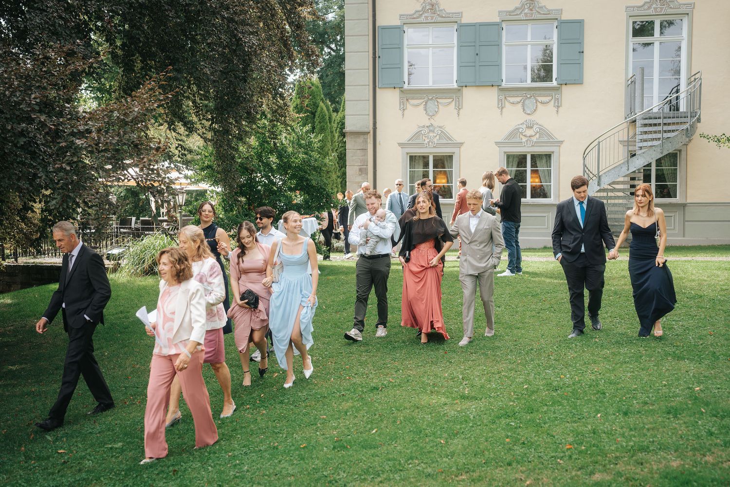 Wedding guests in elegant dresses and suits walk across a sunny green lawn toward a historic pale yellow villa with shuttered windows.