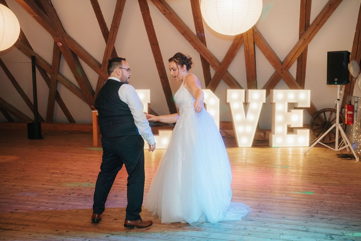 Bride in a white gown and groom in a vest share a first dance on a wooden floor in front of glowing marquee LOVE letters.