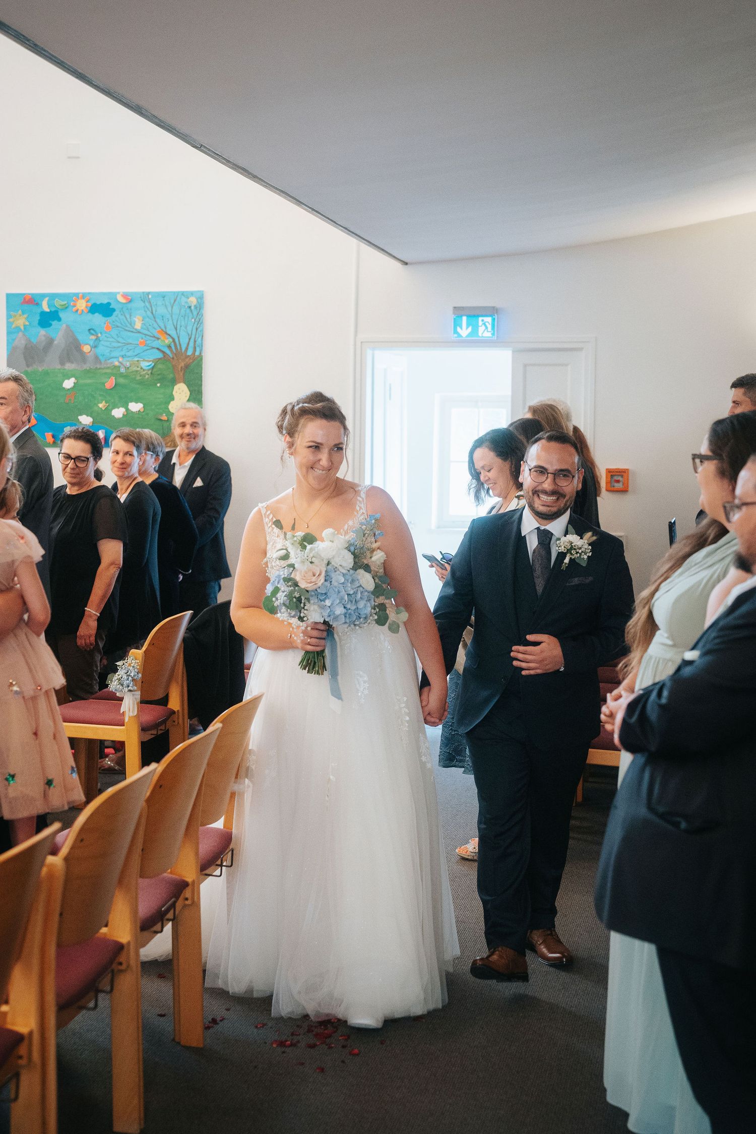 Smiling bride in a white tulle gown holding a blue and white bouquet walks down the chapel aisle beside her groom as guests look on.