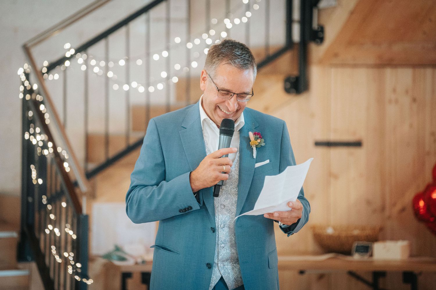 Man in a teal blazer and glasses reads from a paper while speaking into a microphone at a rustic wedding venue with string lights.