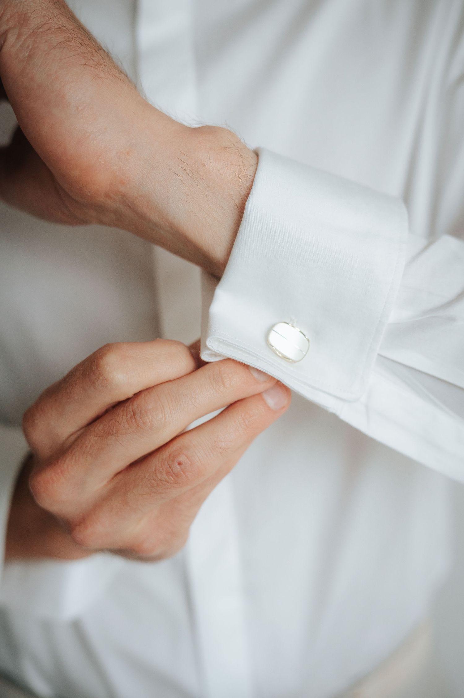 Man in a crisp white dress shirt adjusting a silver oval cufflink on his French cuff sleeve.