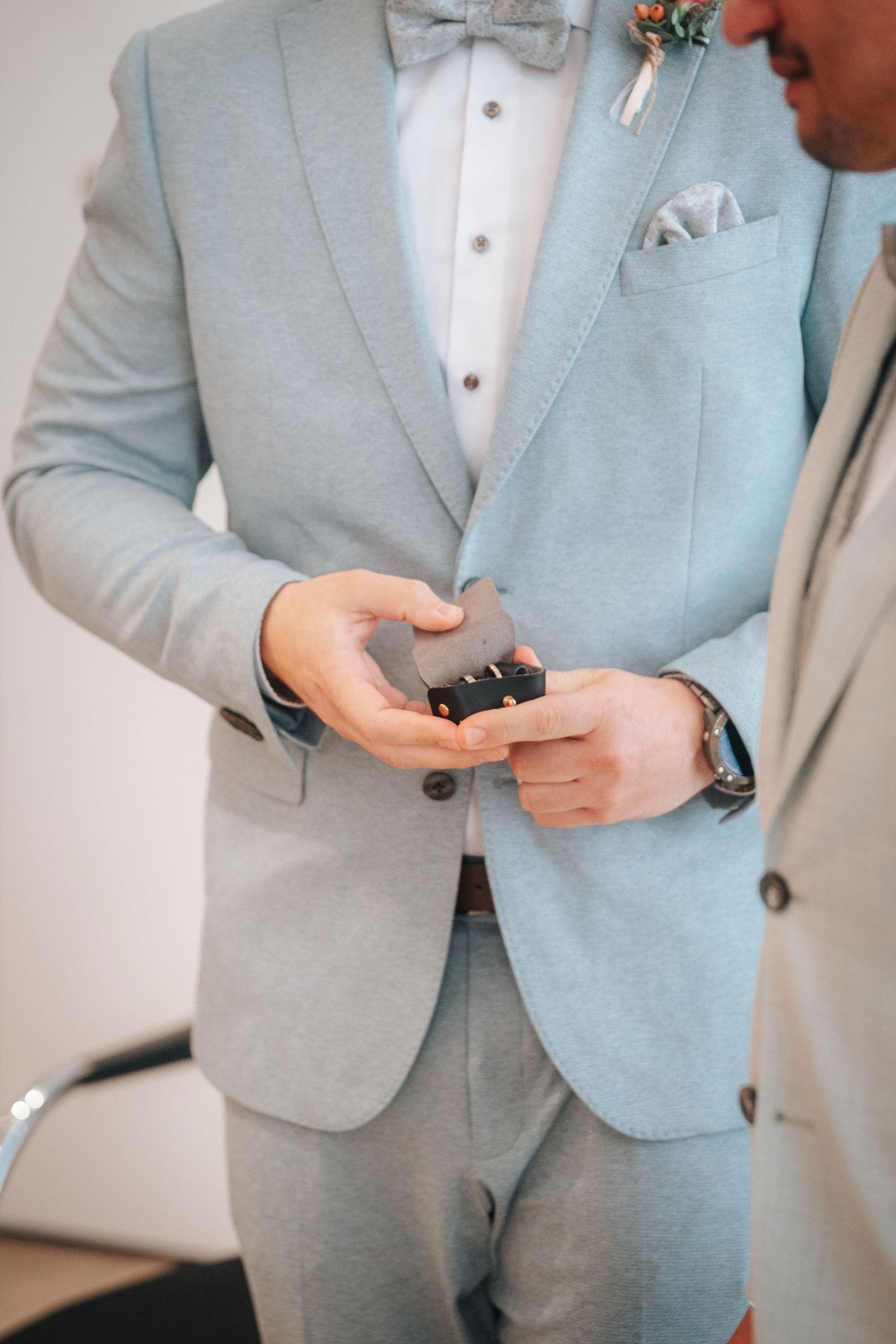 Groom in a light blue suit and bow tie holds a small ring box in his hands during a wedding ceremony.