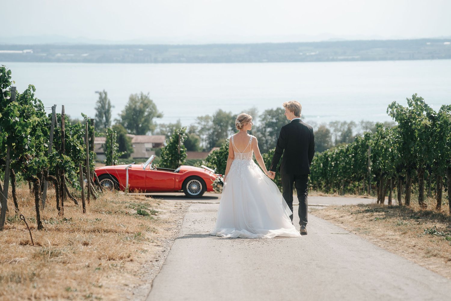 Bride in a white gown and groom in a dark suit walk hand in hand toward a red vintage convertible beside vineyards with a lake view.