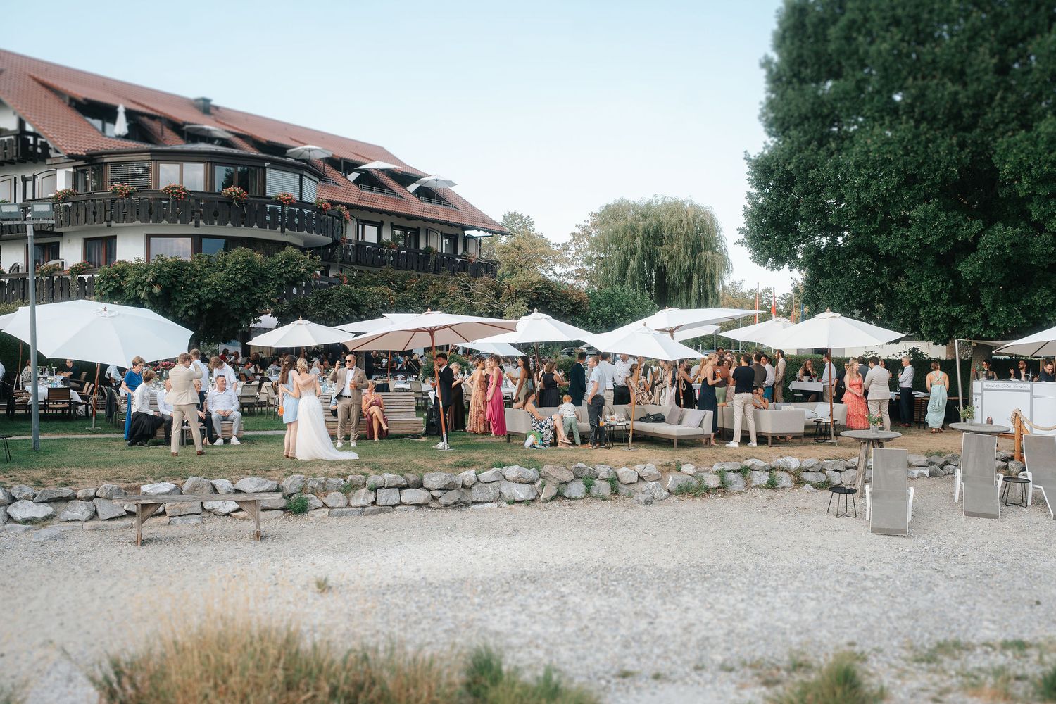 Wedding guests in formal attire gather under white tents on a lakeside lawn near a stone shoreline and a mountain-style lodge.