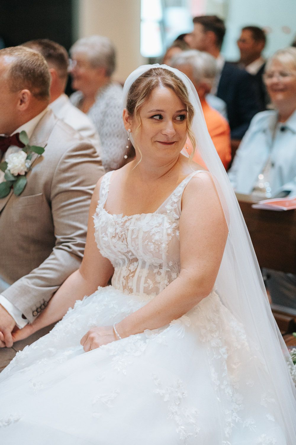A bride wearing a white lace wedding gown and veil sits during a church ceremony with guests in the background.