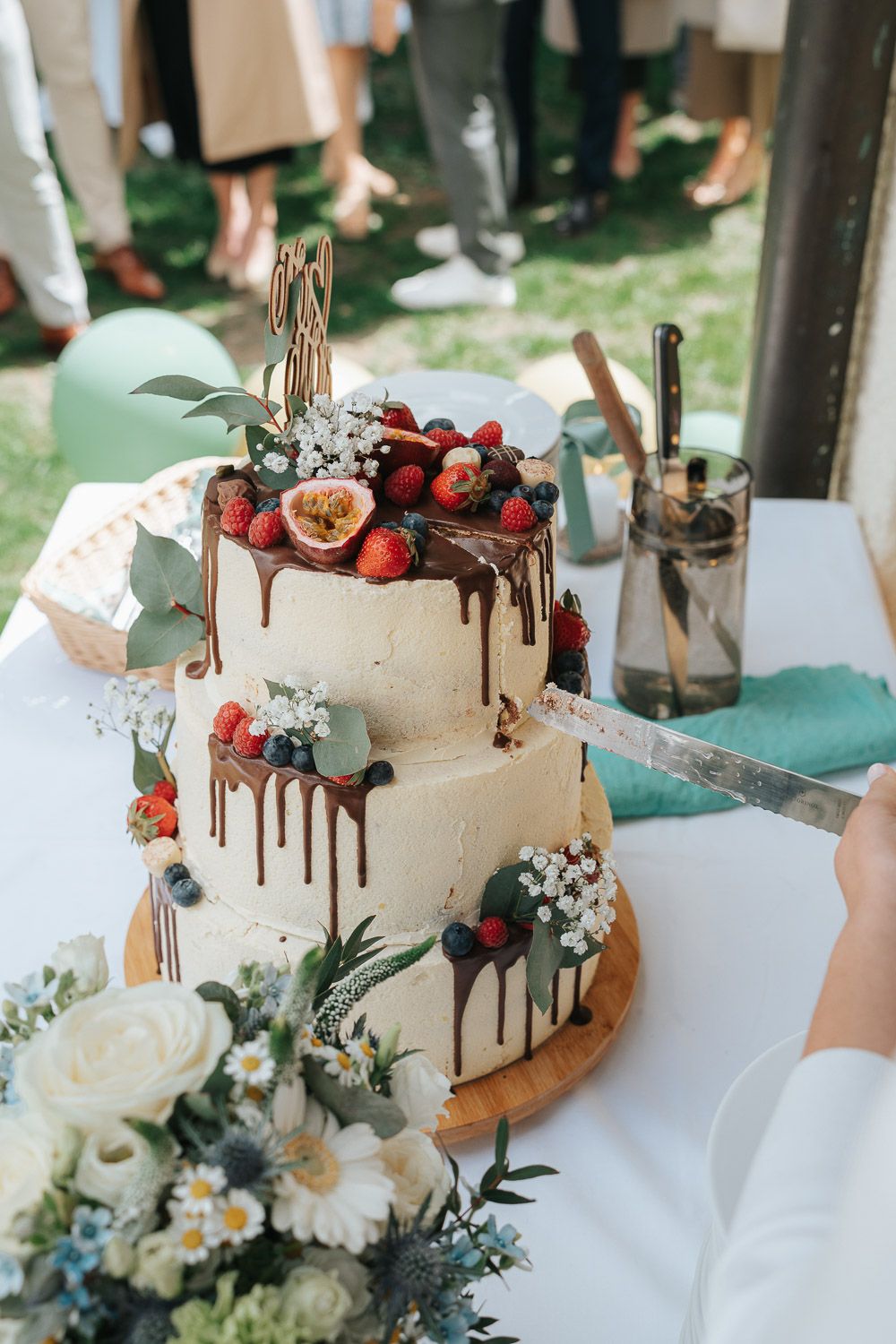 A rustic three-tier wedding cake decorated with chocolate drips, fresh berries, figs and flowers on a wooden cake stand.
