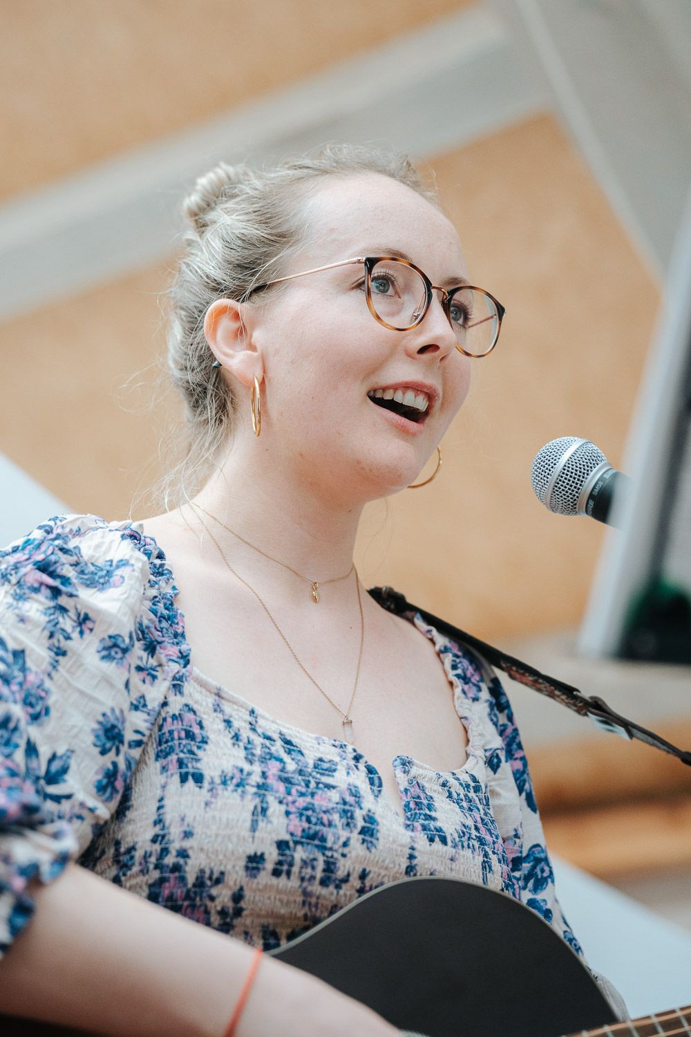 A performer in glasses and a blue floral dress sings into a microphone during a live performance.