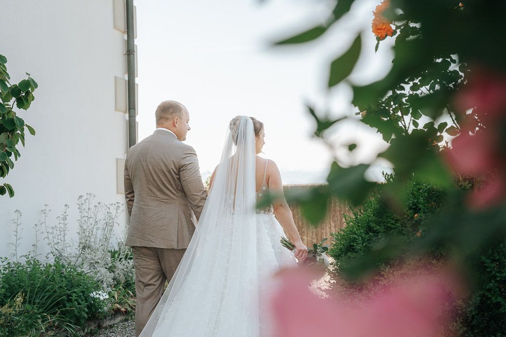 Romantic wedding couple stands together near white wall with flowing veil and garden flowers in soft sunlight.