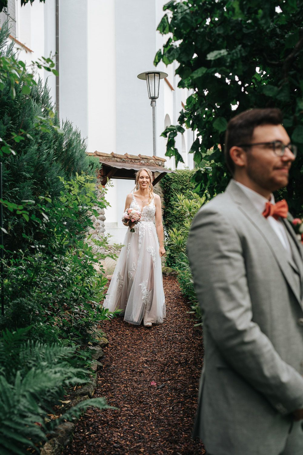 Bride in elegant lace gown stands on garden path behind groom in gray suit during outdoor wedding first look moment.