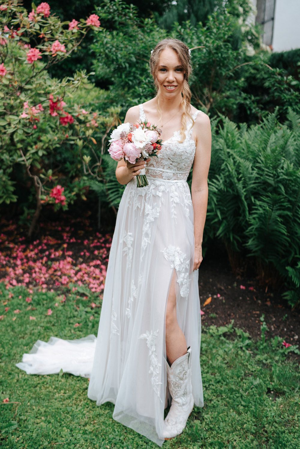 A stunning bride wearing a white lace dress with floral appliques poses outdoors in a garden setting with blooming flowers.