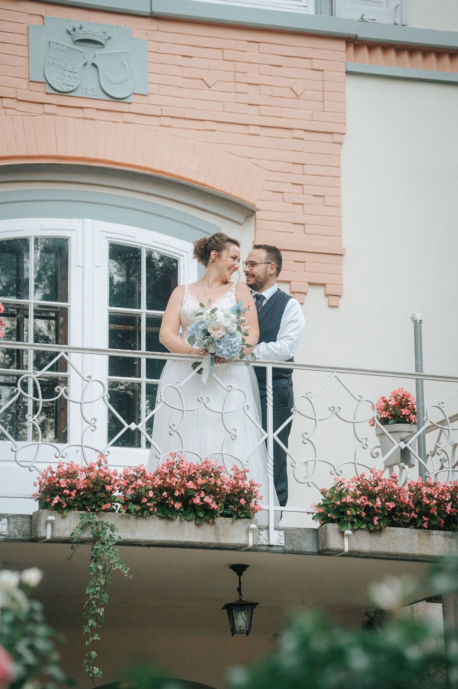 Bride in a white gown holding a bouquet and groom in a vest share a smiling moment on a balcony with pink flower boxes.