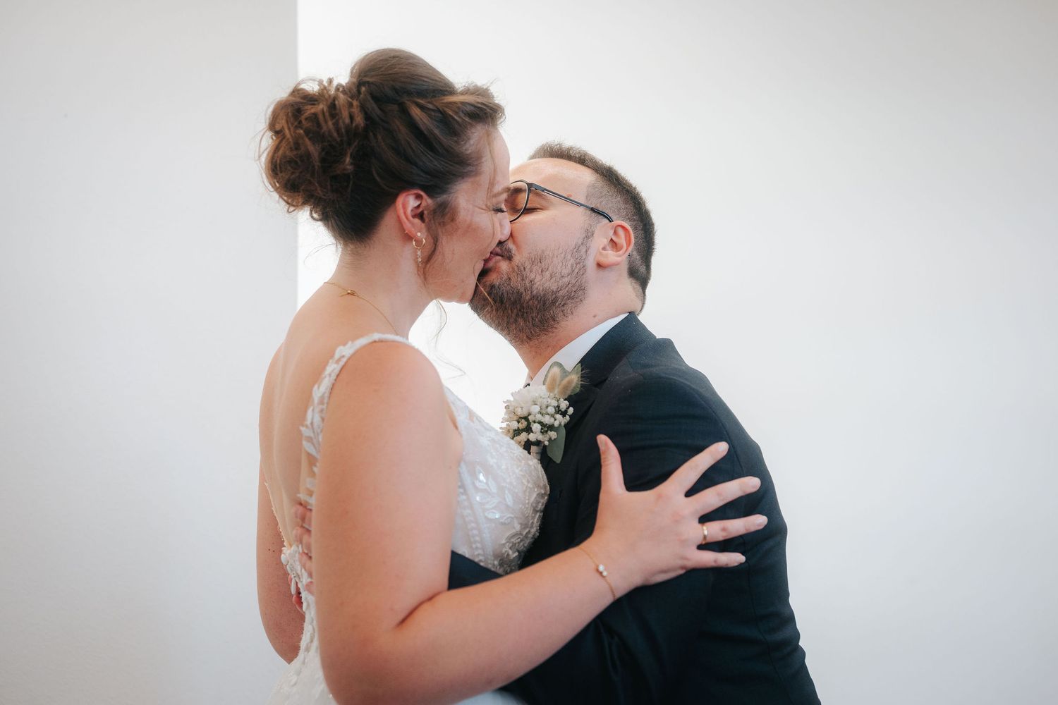 Bride in a white lace wedding dress and groom in a dark suit with a boutonniere share a kiss during their wedding ceremony.