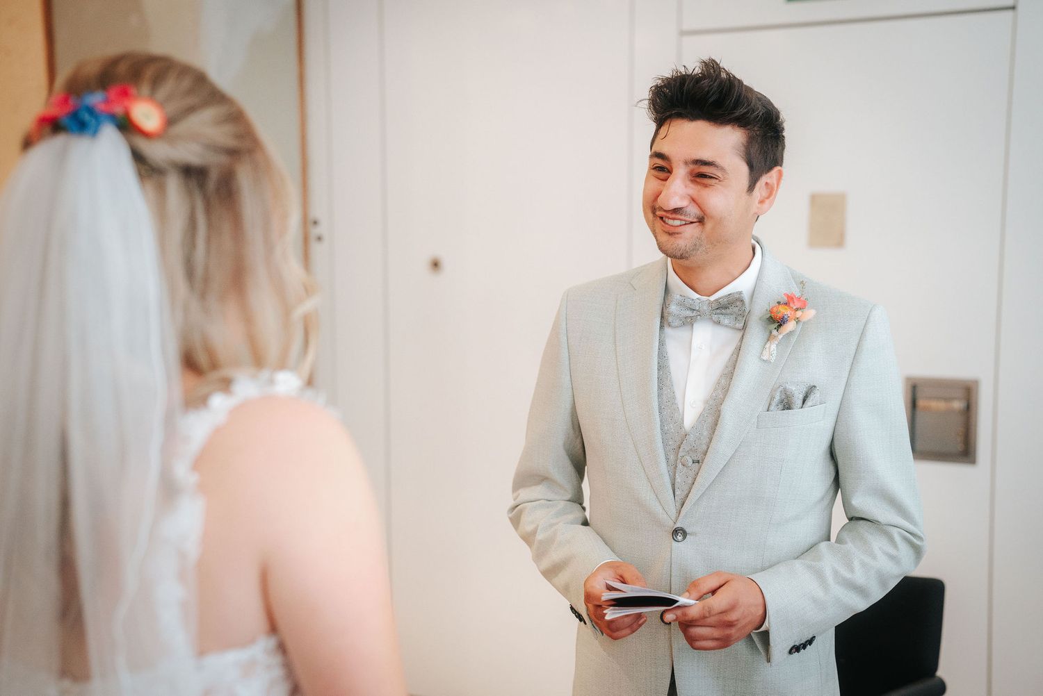 Smiling groom in a light gray suit and bow tie reads his vows from a small card to a bride wearing a veil and floral hair accessory.