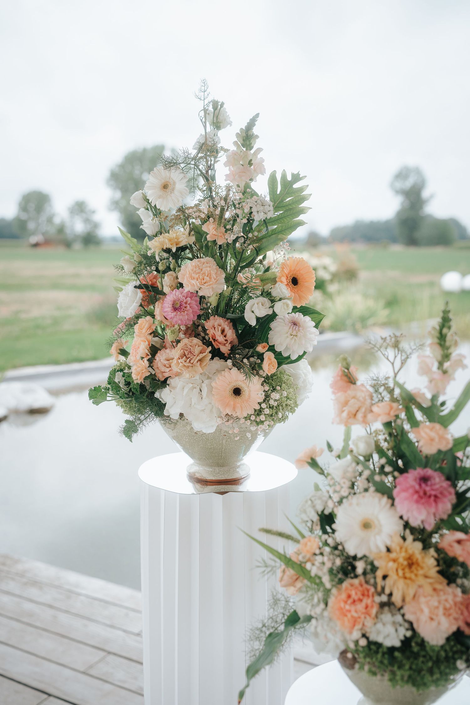Pastel peach, pink and white floral arrangements in urn vases on white pedestals beside a calm pond, ideal for outdoor wedding decor.