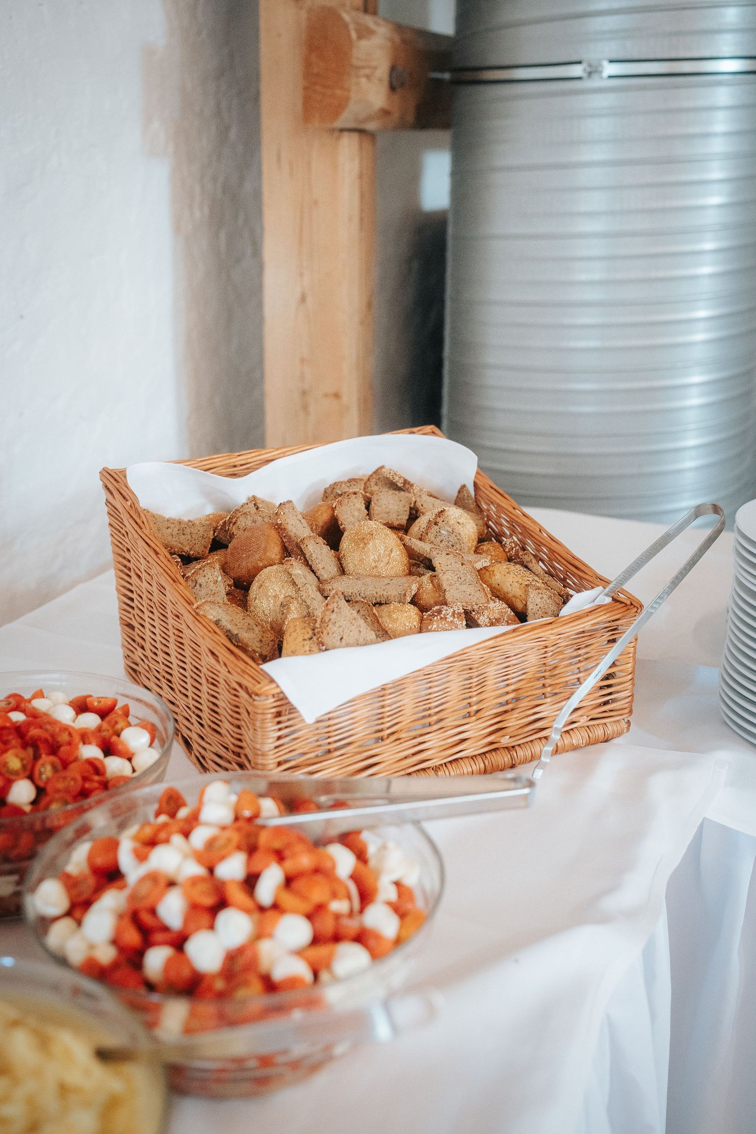 Buffet table with a wicker basket of sliced rustic bread, tongs, and glass bowls of cherry tomato and mozzarella salad.