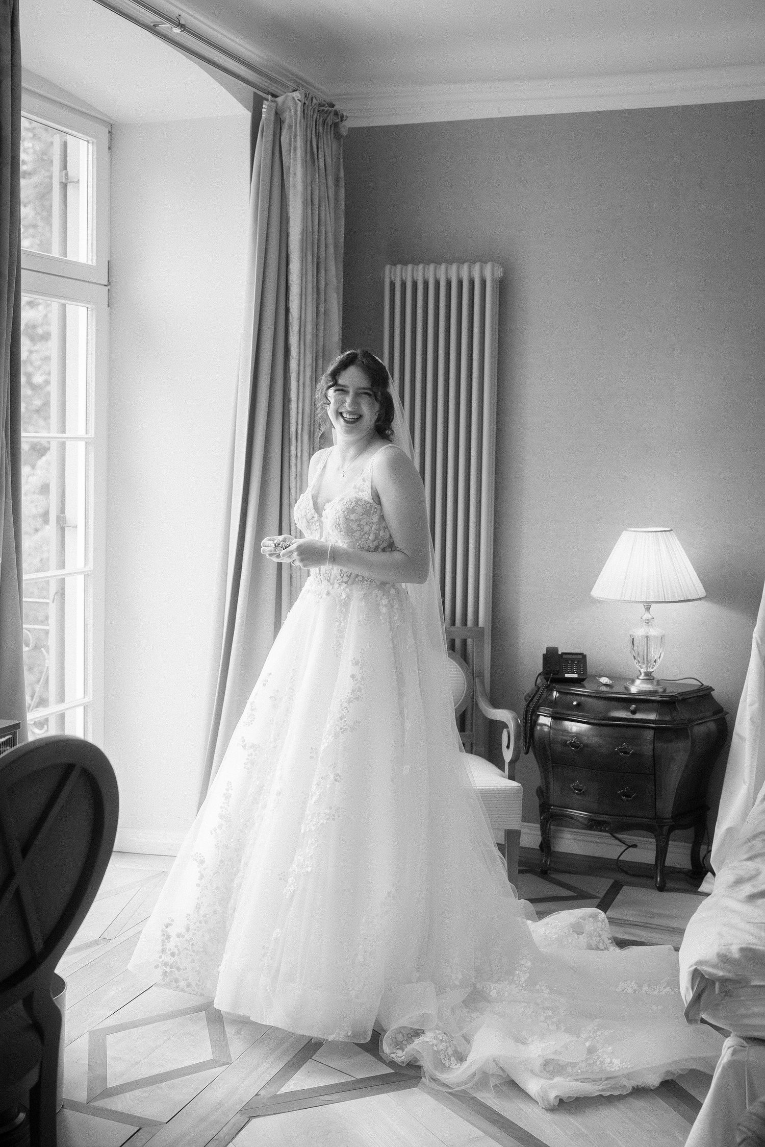 Smiling bride in a lace A-line wedding gown with a long train stands beside tall windows in an elegant room, in black and white.