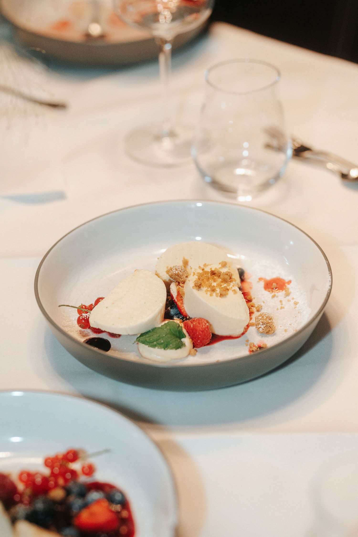 Elegant plated dessert with creamy white quenelles, fresh berries, mint and crumble in a gray bowl on a restaurant table.