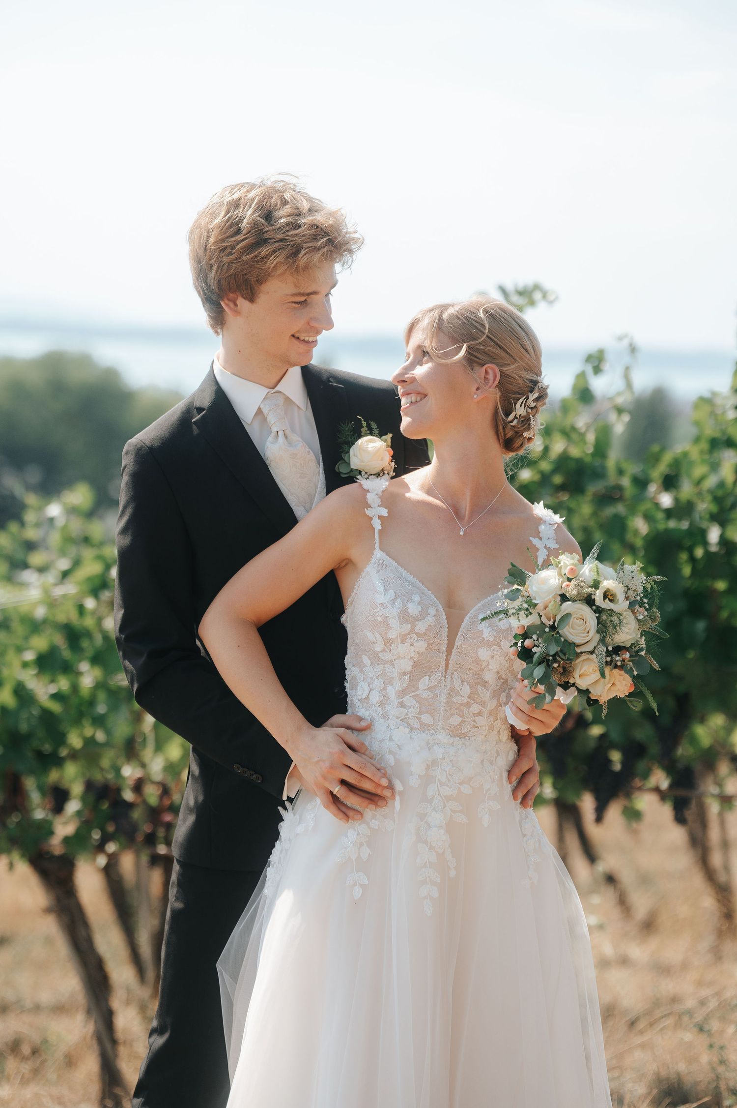 Smiling bride in a lace wedding gown holding a bouquet embraces her groom in a black suit with a boutonniere among sunlit vineyard rows.