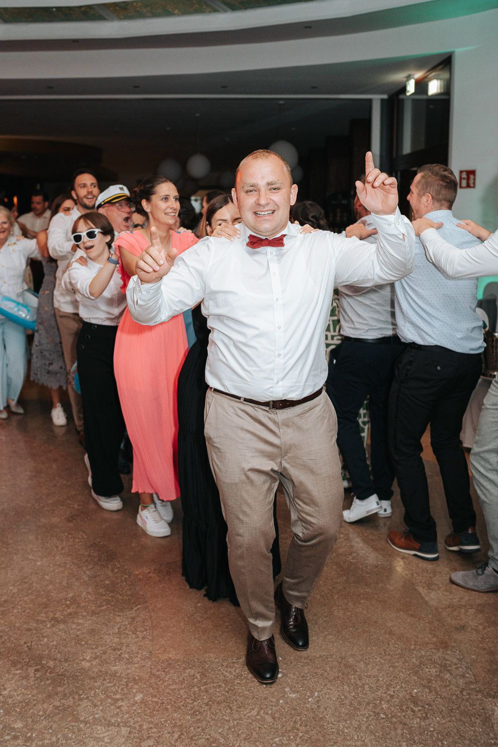 Wedding guests dancing in line at a reception, with dancers wearing formal attire and having fun on the dance floor.