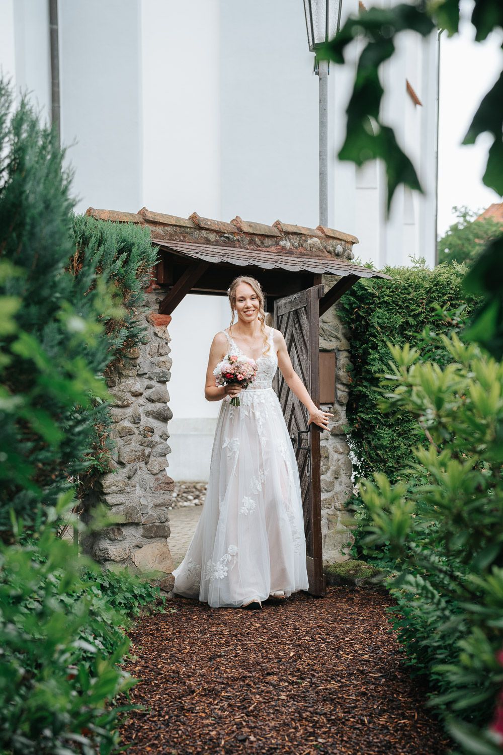 A bride in a white lace wedding gown stands in a rustic stone doorway surrounded by green foliage.