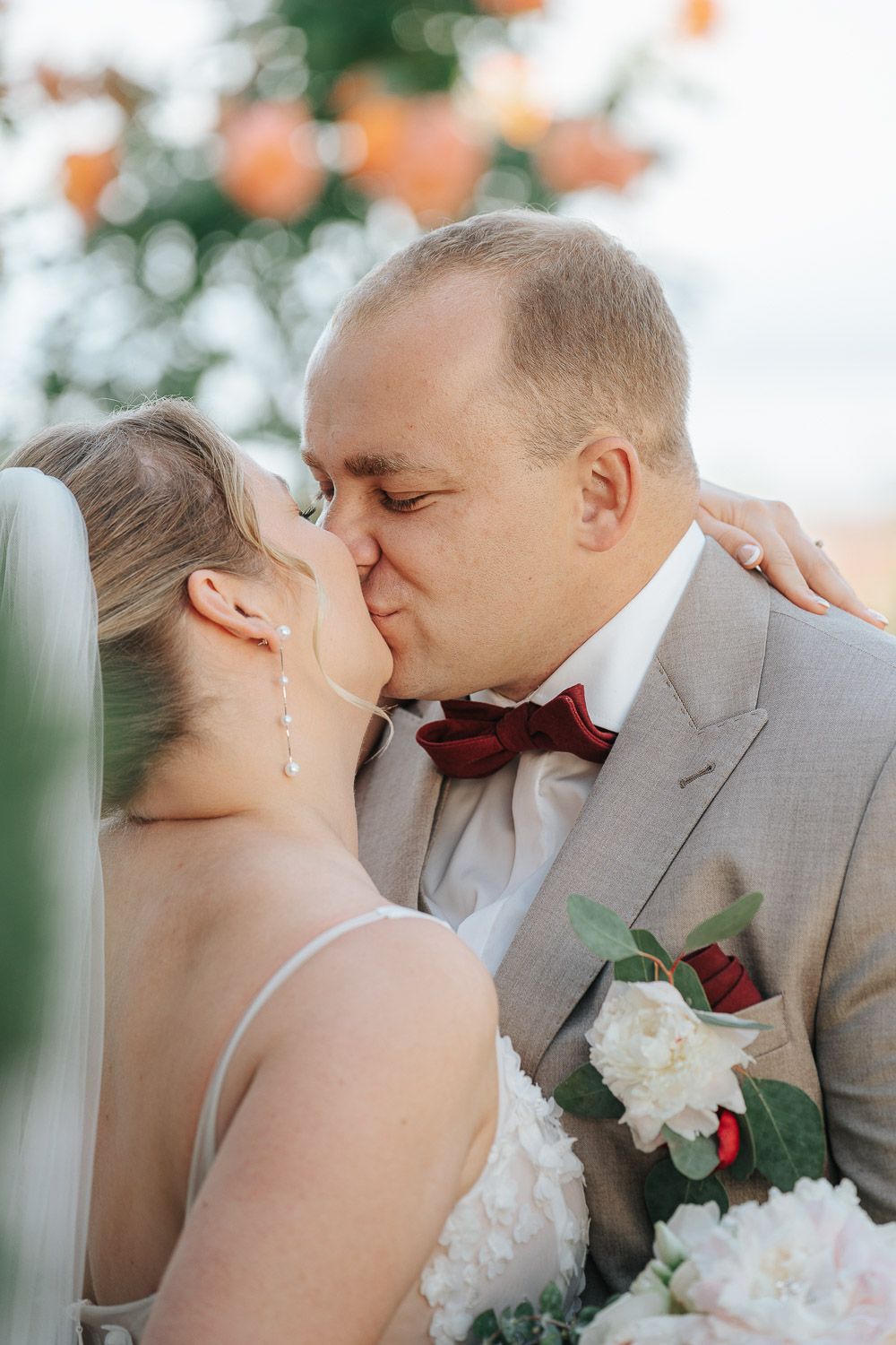 Bride in white gown and groom in gray suit with red bowtie share tender kisses during their wedding portraits.