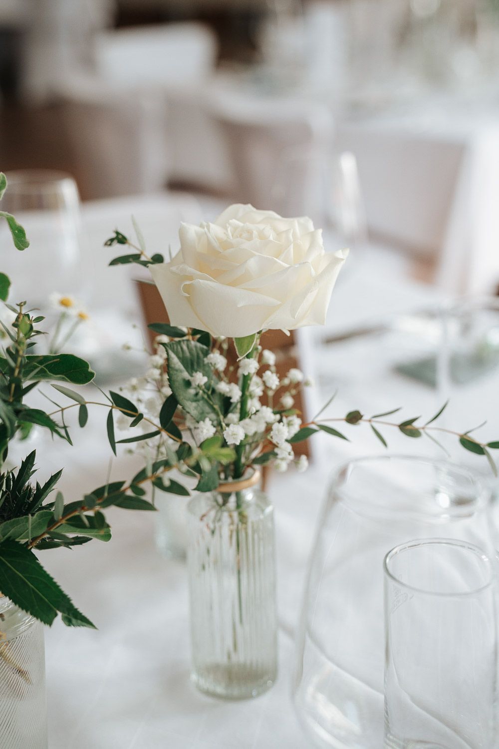 A delicate white rose in a glass vase with eucalyptus and greenery on an elegant white tablecloth for a wedding reception.