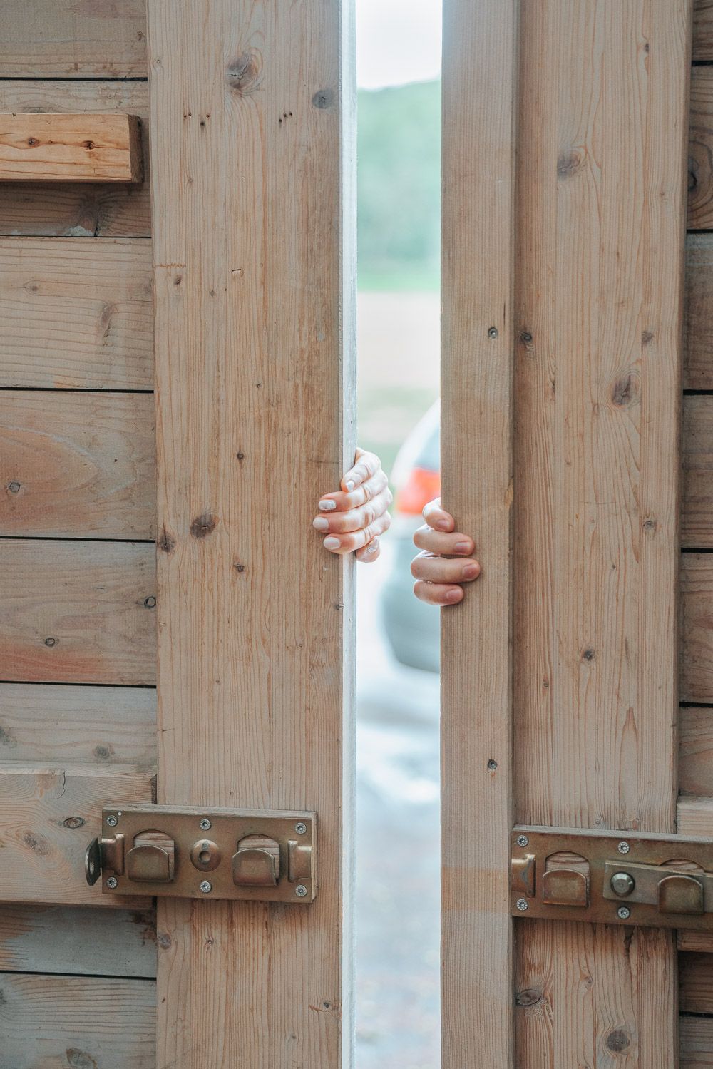 Two hands reaching through a gap between wooden boards with metal latches, creating an eerie or trapped atmosphere.