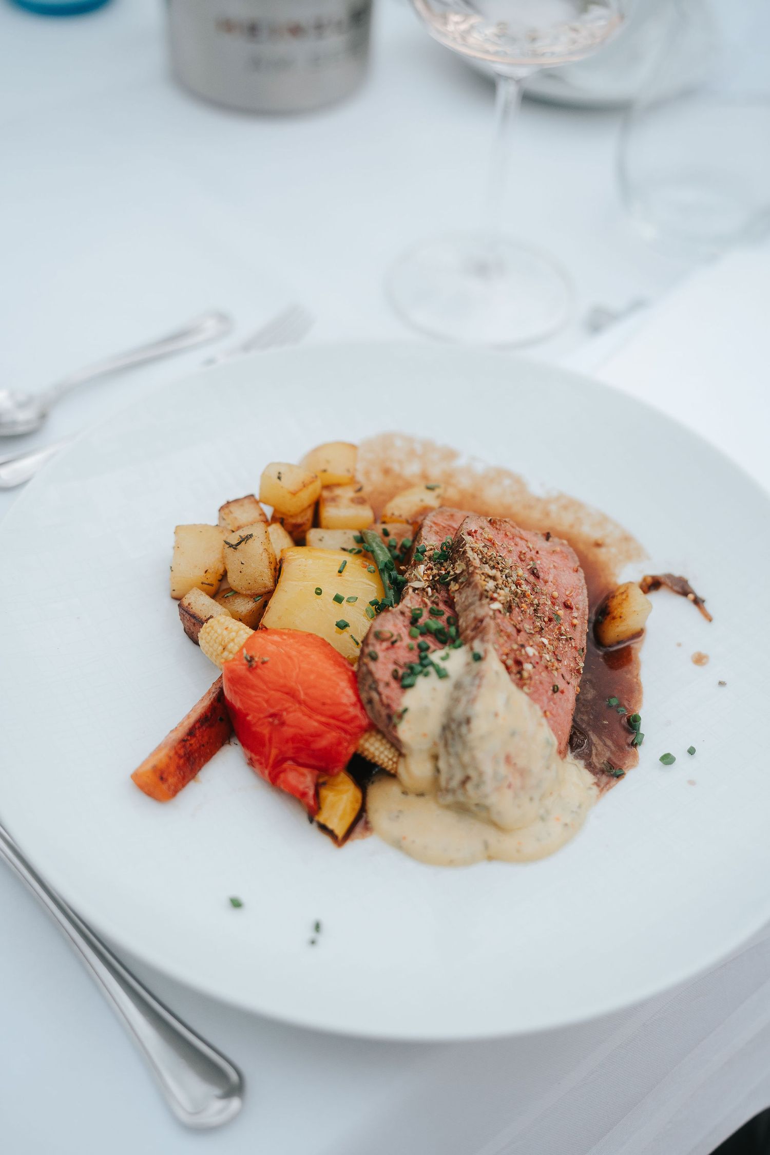 Plated roasted meat with creamy sauce, glazed carrots, diced potatoes and herbs on a white plate at an elegant table setting.