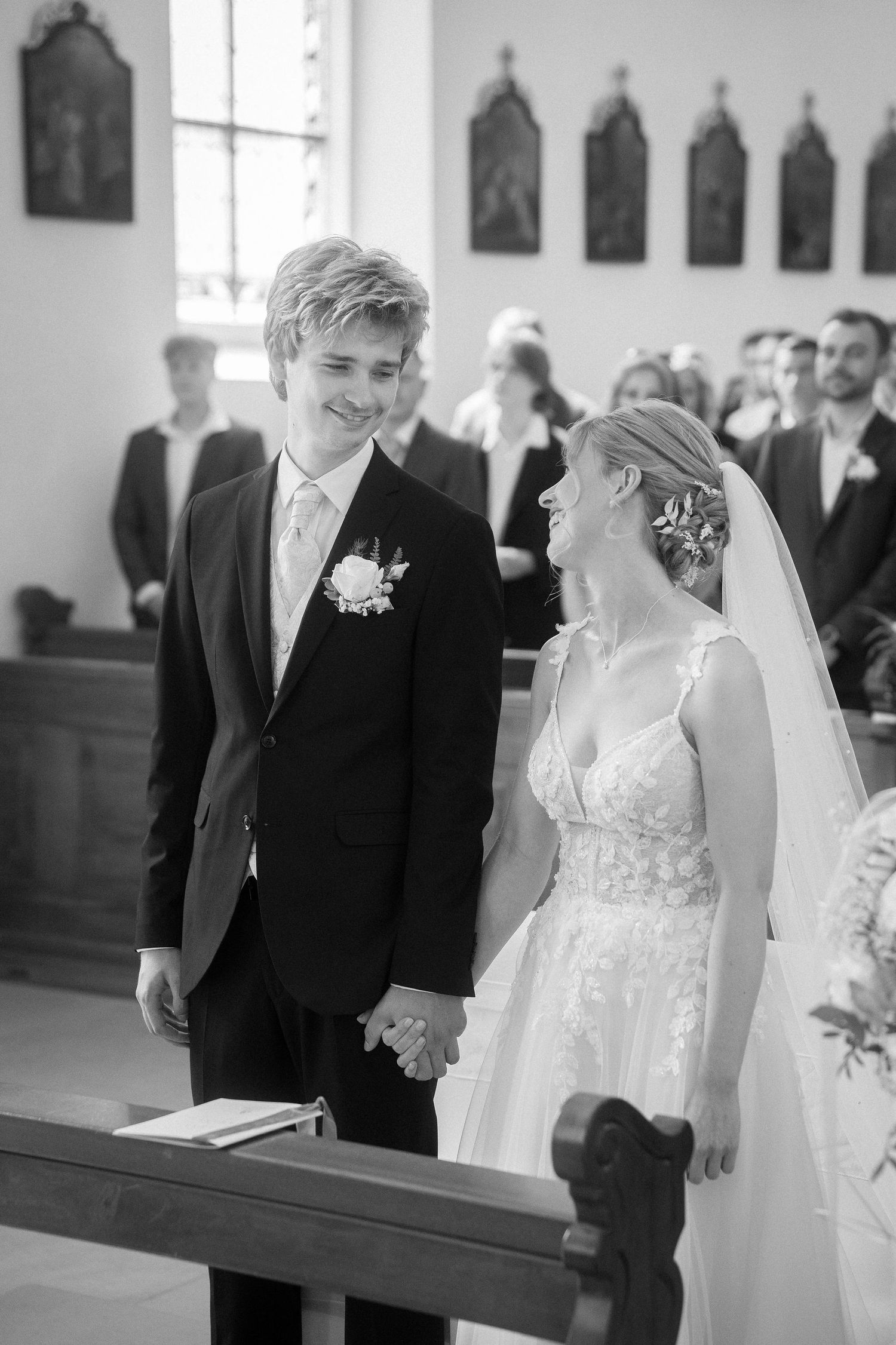 Smiling groom in a dark suit holds hands with a bride in a lace gown and veil during a wedding ceremony in a church.
