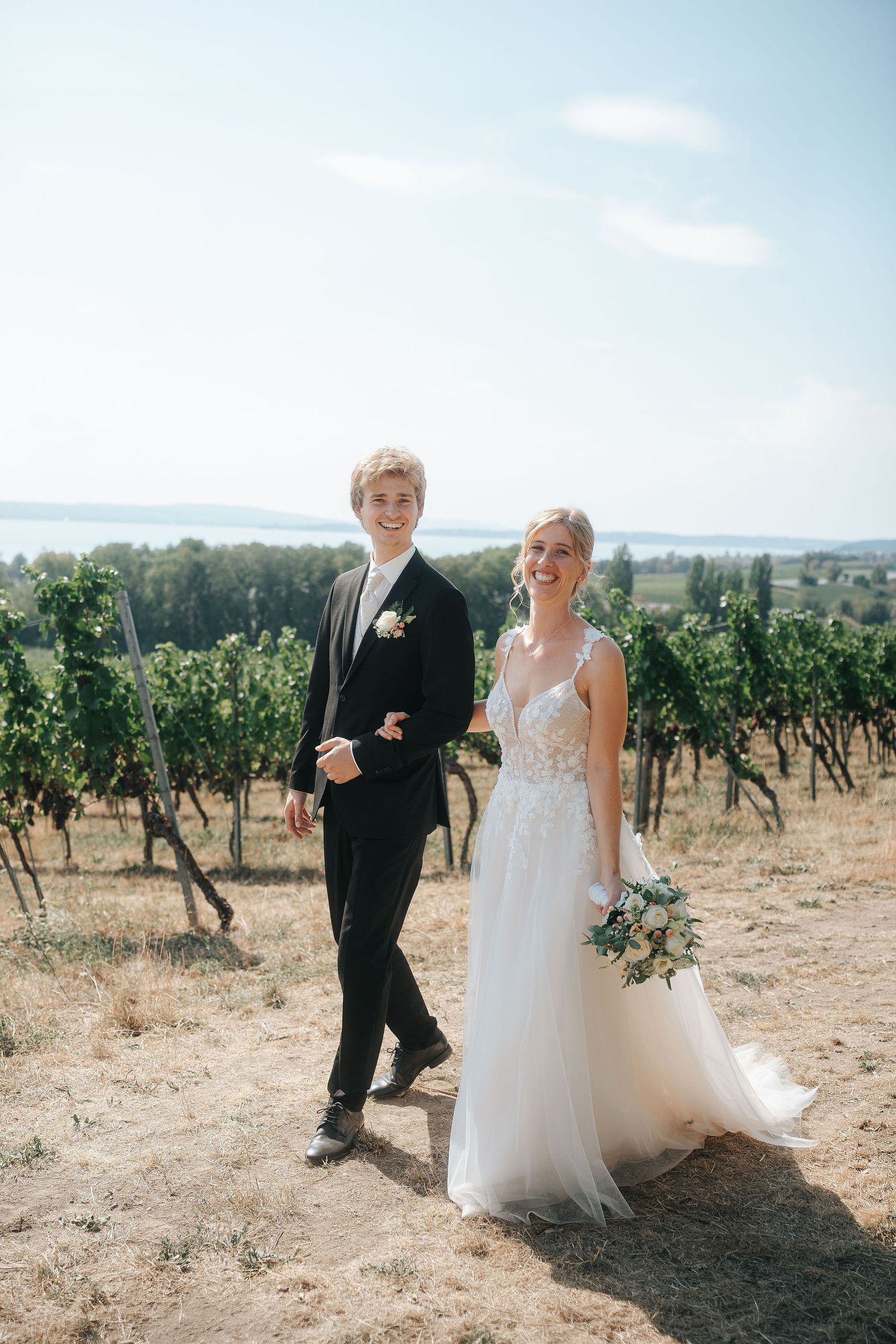 Smiling bride in a lace wedding gown holding a bouquet walks arm in arm with the groom in a black suit through a sunny vineyard.