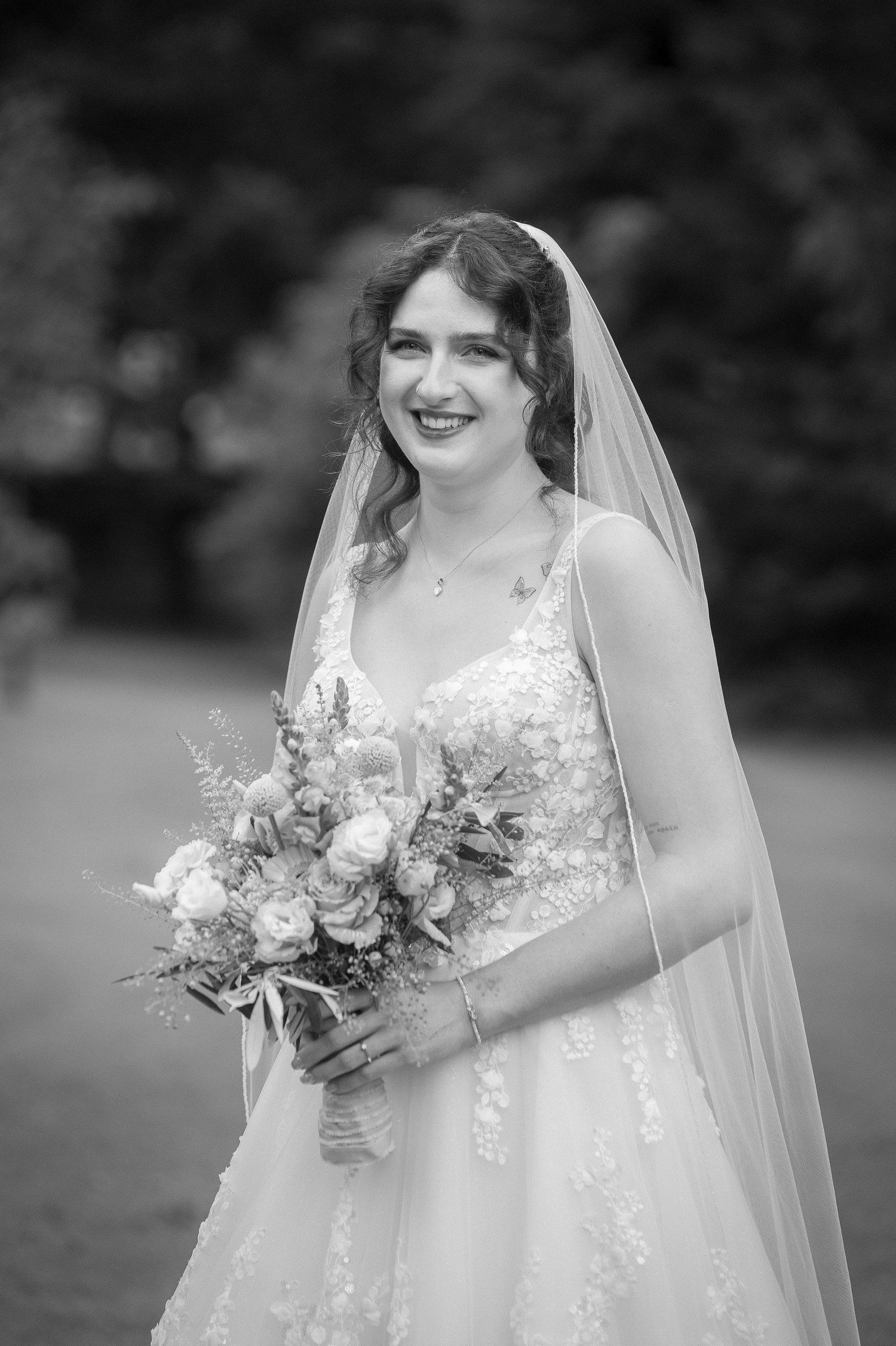 Black and white portrait of a smiling bride in a lace gown and long veil holding a bouquet outdoors.