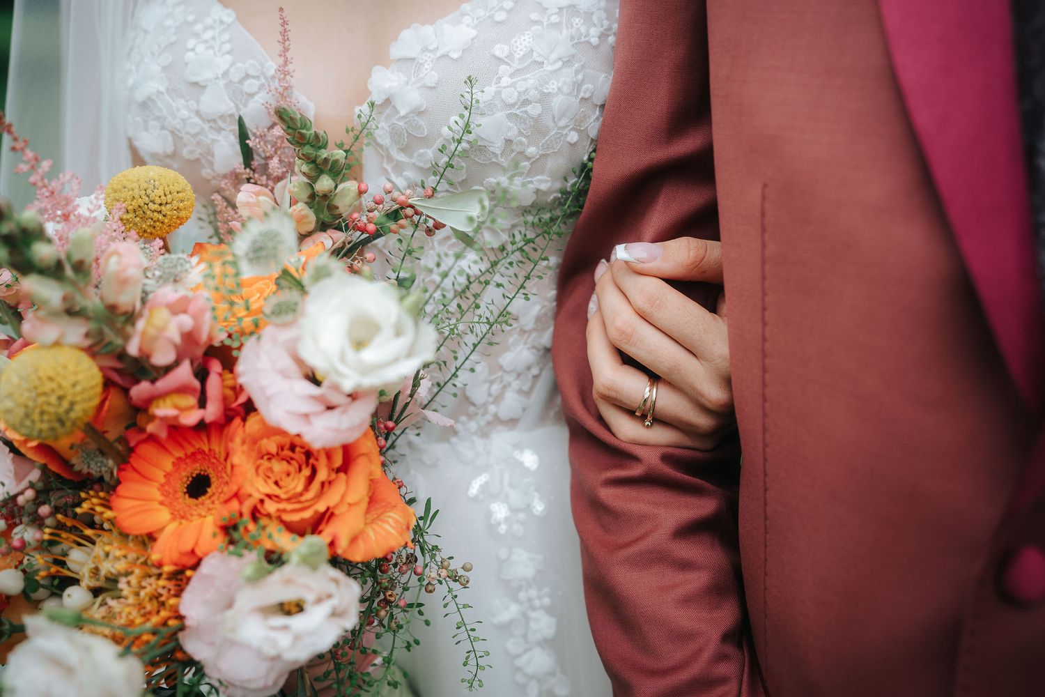 Bride in a lace wedding gown holds an orange and blush bouquet while resting her ringed hand on the groom's rust-colored suit sleeve.