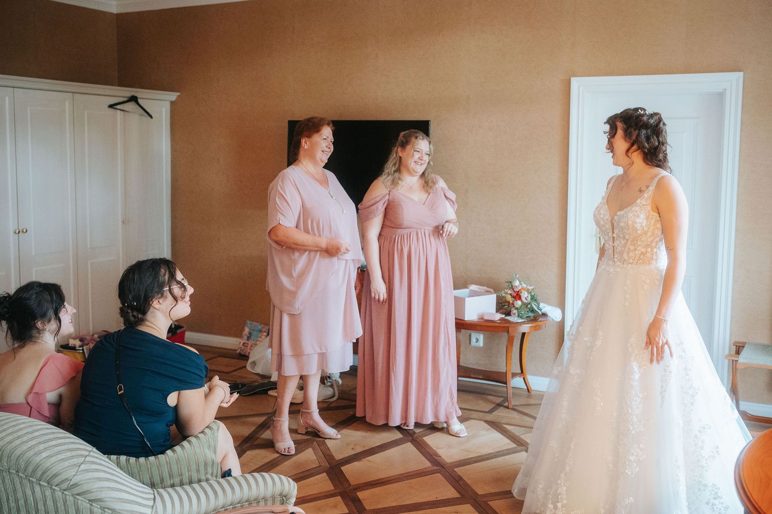 Bride in a white lace wedding gown smiles as bridesmaids in pink dresses and seated guests look on in an elegant getting-ready room.